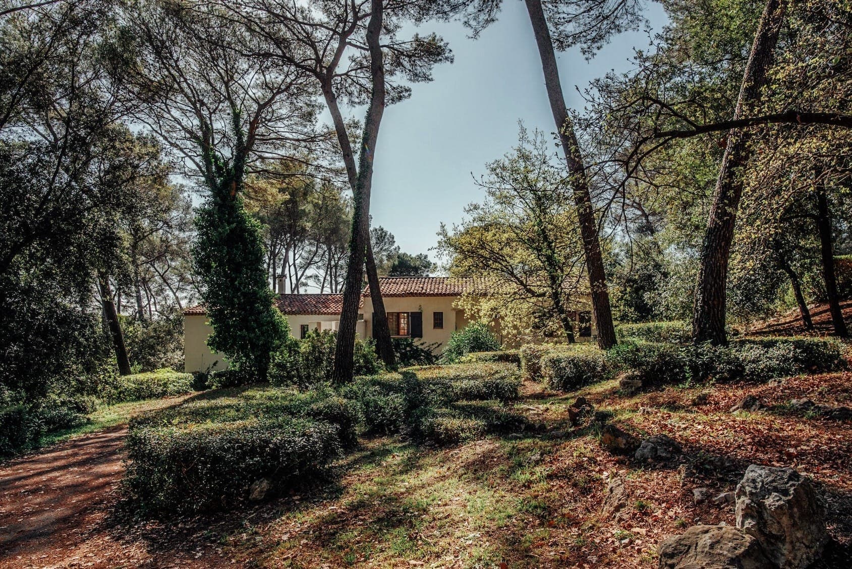 Une petite maison au toit de tuiles rouges est nichée au milieu de grands pins et d'une végétation luxuriante. Le soleil filtre à travers les arbres, projetant des ombres tachetées sur le sol. Un sentier mène à la maison, créant une scène sereine et boisée.