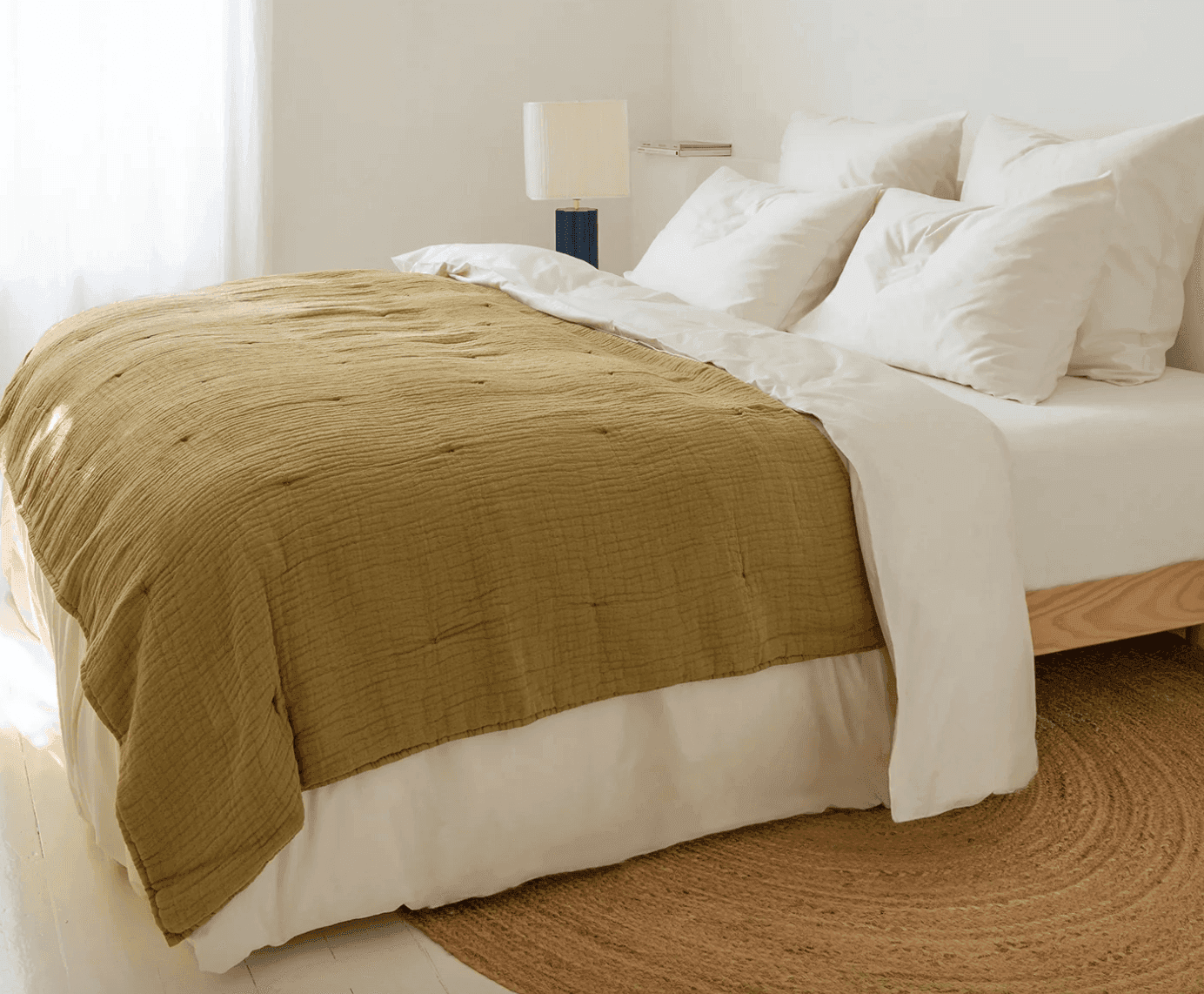 Un lit bien fait avec des oreillers et des draps blancs et un edredon en satin vert olive. Une petite lampe et un tapis rond tissé se trouvent à proximité, contre un mur blanc et une pièce doucement éclairée.