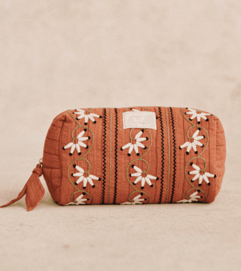 Pochette cylindrique marron avec broderie florale en blanc, rose et vert. Elle est dotée d'une fermeture éclair avec tirette en tissu et d'une petite étiquette sur un côté. Le fond est un tissu doux et neutre.