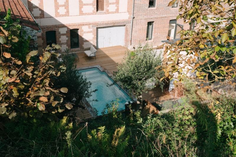 A small rectangular pool with clear water sits on a wooden deck beside a brick building, surrounded by leafy trees and greenery on a sunny day. An empty chair is placed near the pool.