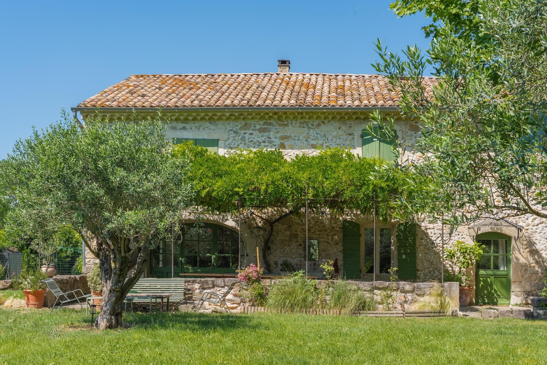 A rustic stone house with a tiled roof and green shutters, surrounded by lush trees, vines, and a grassy yard under a clear blue sky. A shaded patio with plants and outdoor furniture is visible in front.