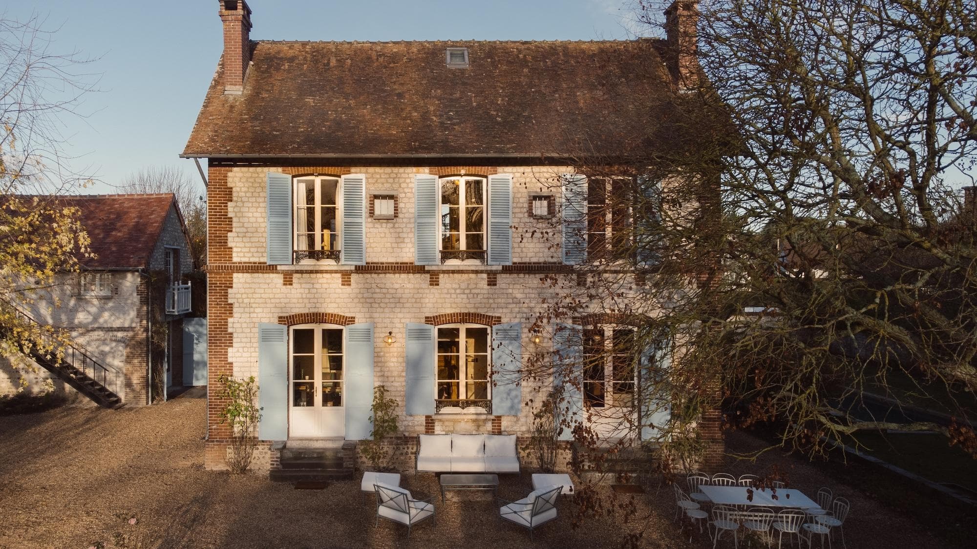 A two-story brick house with blue shutters, large windows, and outdoor seating in the front yard. Trees and patio furniture are visible, and the scene is lit by warm sunlight.