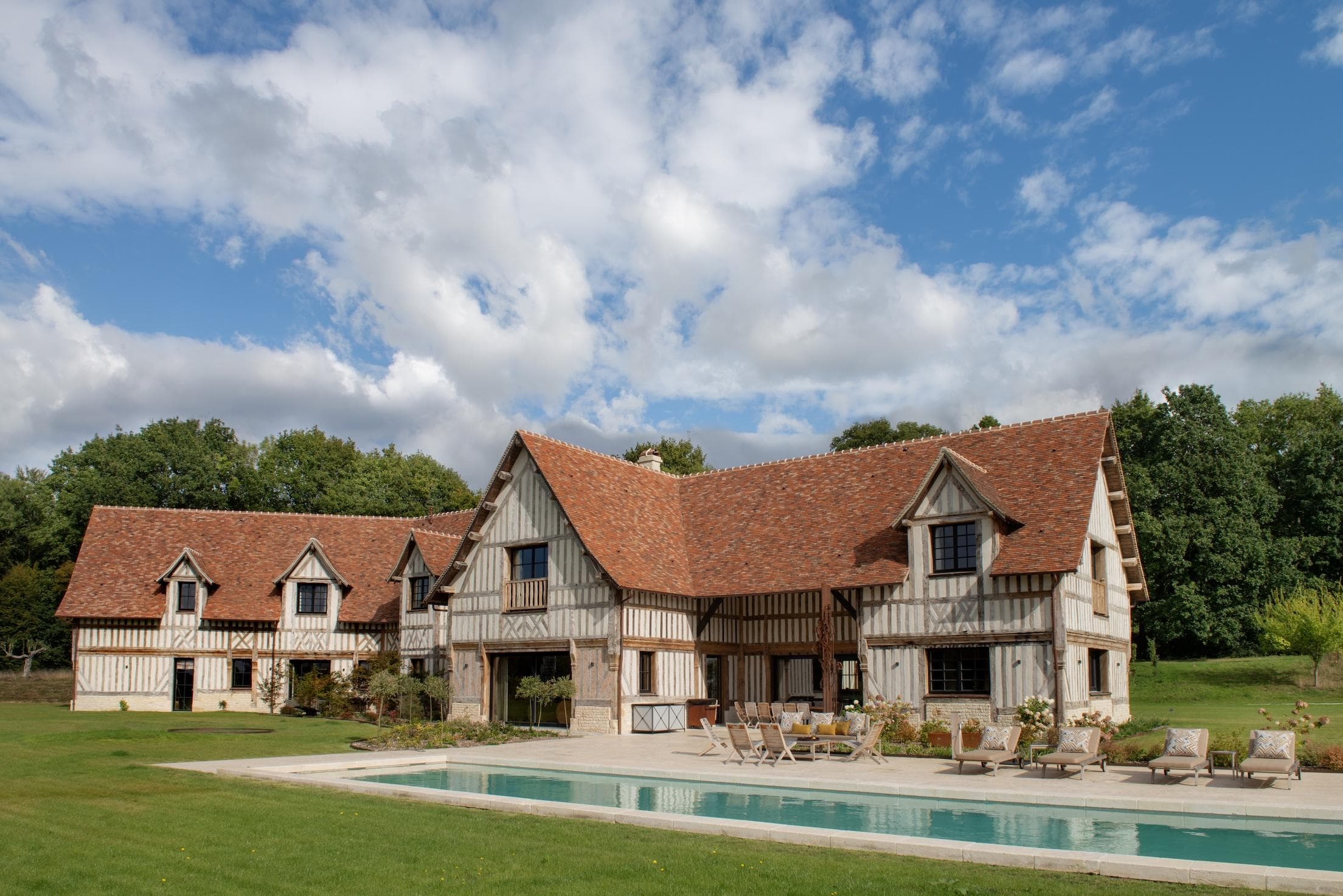 A large, traditional half-timbered house with a red-tiled roof sits next to a rectangular swimming pool, surrounded by green lawn and trees under a partly cloudy sky.
