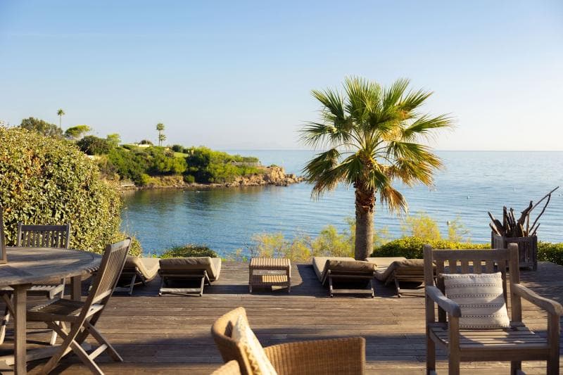 A sunny deck with wooden chairs and tables overlooks the calm sea, with lounge chairs facing the water, a palm tree, and green coastal foliage in the background.