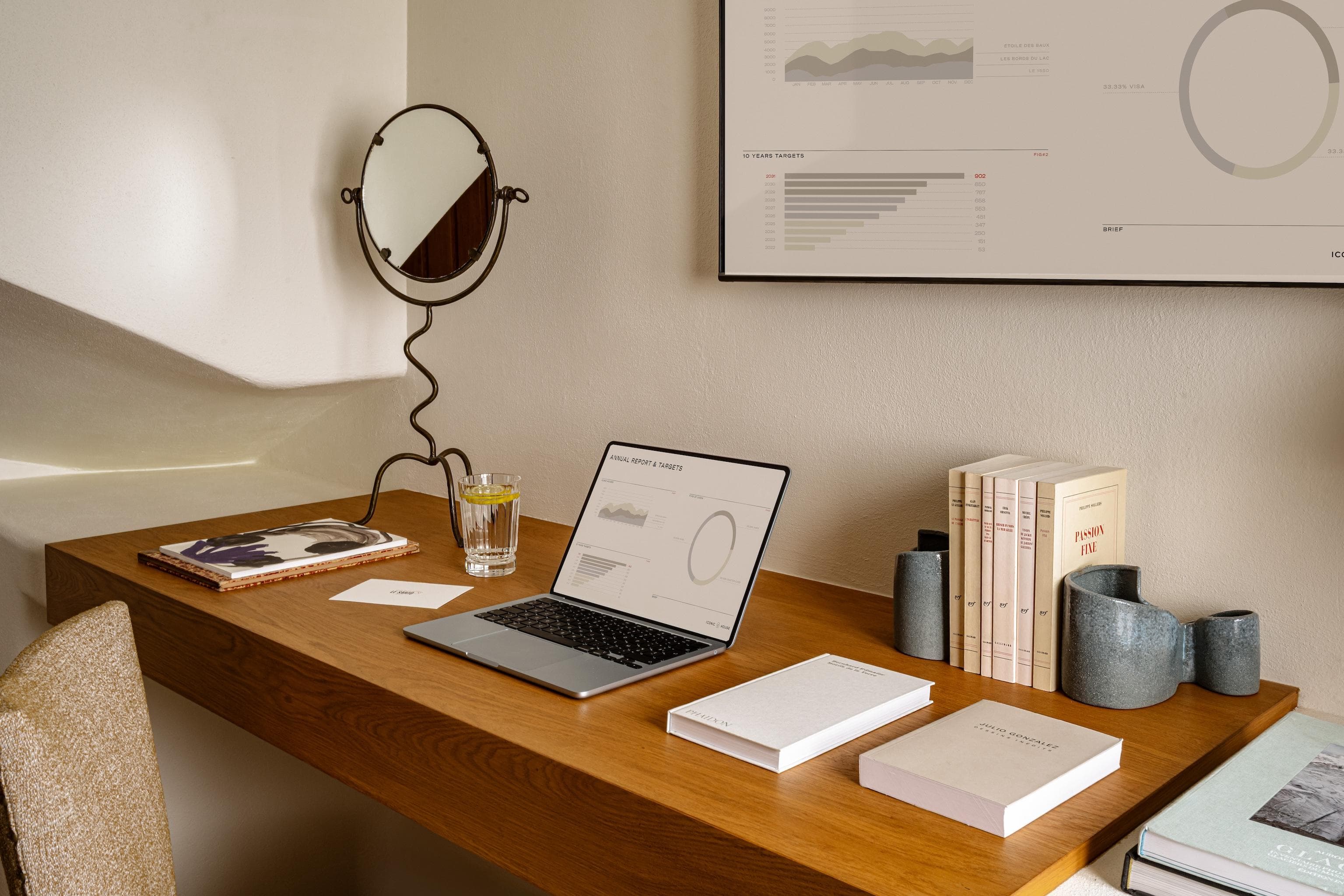 A minimalist wooden desk with a laptop, notebooks, books, a glass of water, a decorative mirror, and a ceramic vase. Modern charts are displayed on the wall above the workspace.
