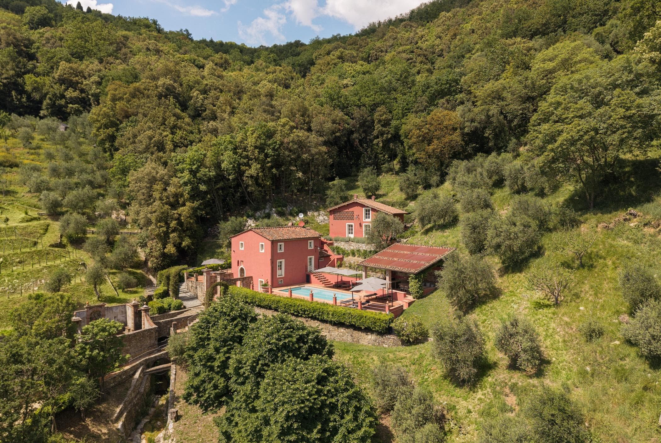 Aerial view of a red villa with a swimming pool, patios, and outbuildings nestled among green trees and hills in a lush, rural landscape.