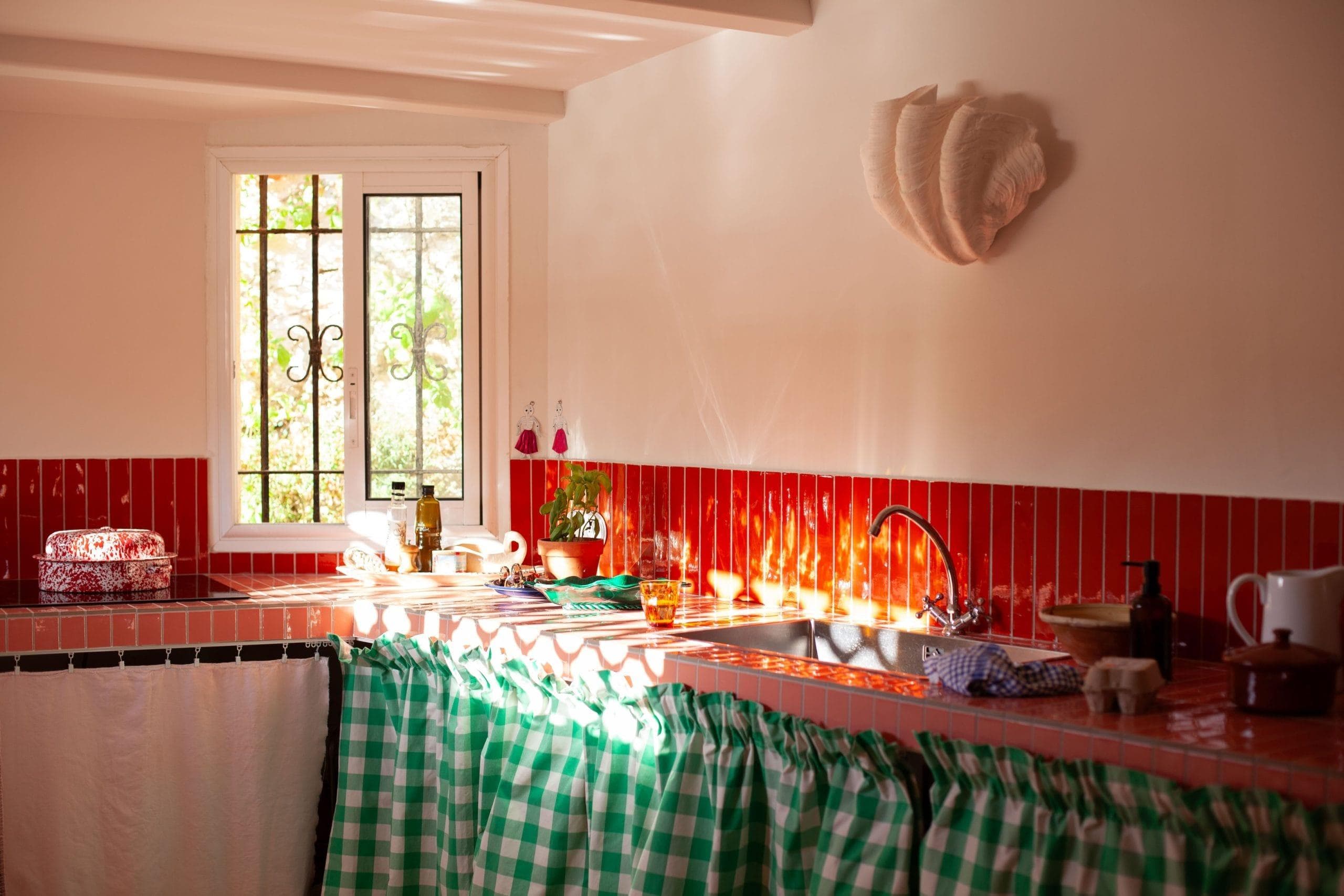 A cozy kitchen reminiscent of The Cabanon des Goudes features a red tiled backsplash and a window letting in sunlight. Green and white checkered curtains hang under the countertop, while plants and kitchen items rest alongside light creating patterns on the walls.