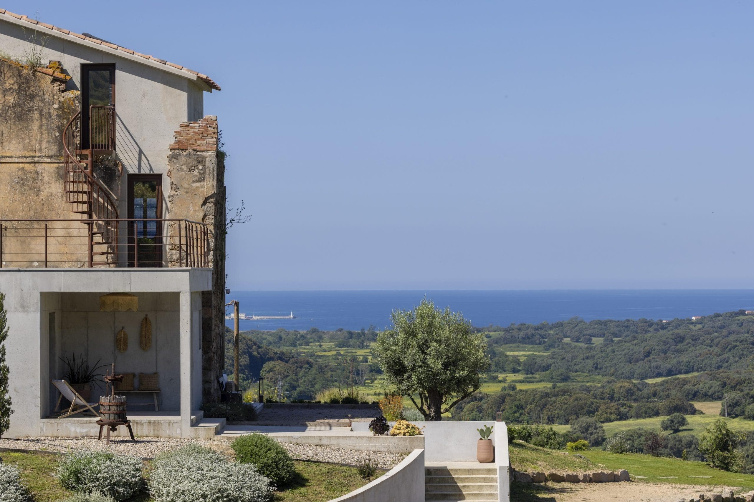 A rustic house with weathered walls and an outdoor terrace overlooks a lush green landscape and the blue sea under a clear sky. A staircase leads to a small garden with a potted plant in the foreground.