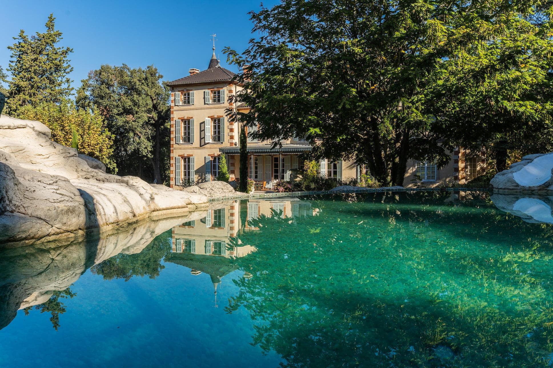 Une belle maison à plusieurs étages, ornée de grandes fenêtres et entourée d'une verdure luxuriante, se reflète dans une piscine bleu clair. Des rochers et des arbres encadrent la piscine, créant un cadre serein et pittoresque par une journée ensoleillée.
