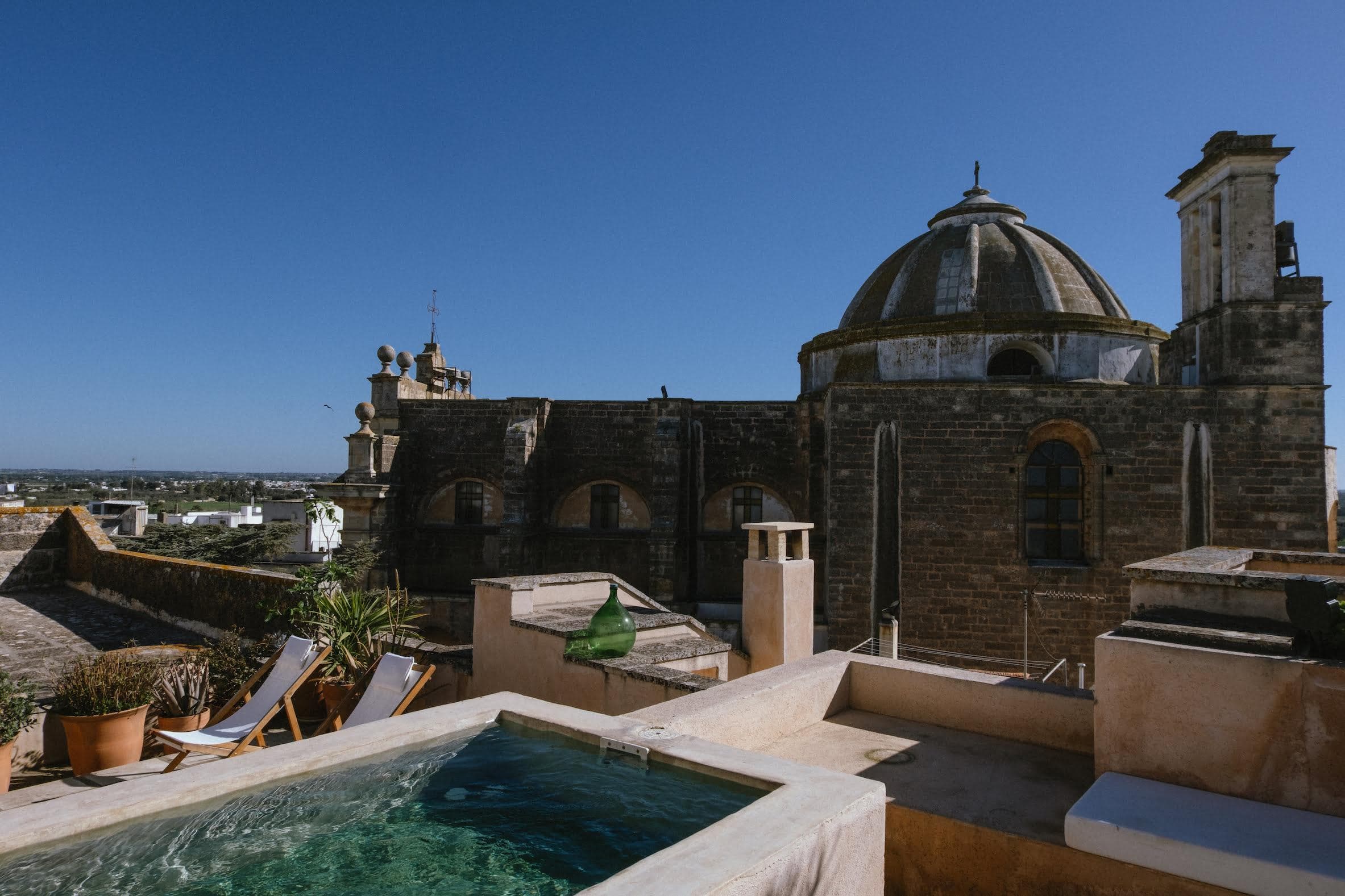 A rooftop scene with a small pool in the foreground, bordered by potted plants and lounge chairs, evokes the luxury of Puglia's finest villas. In the background, an old stone building with arched windows and a large dome stands under a clear blue sky, perfect for a holiday retreat.