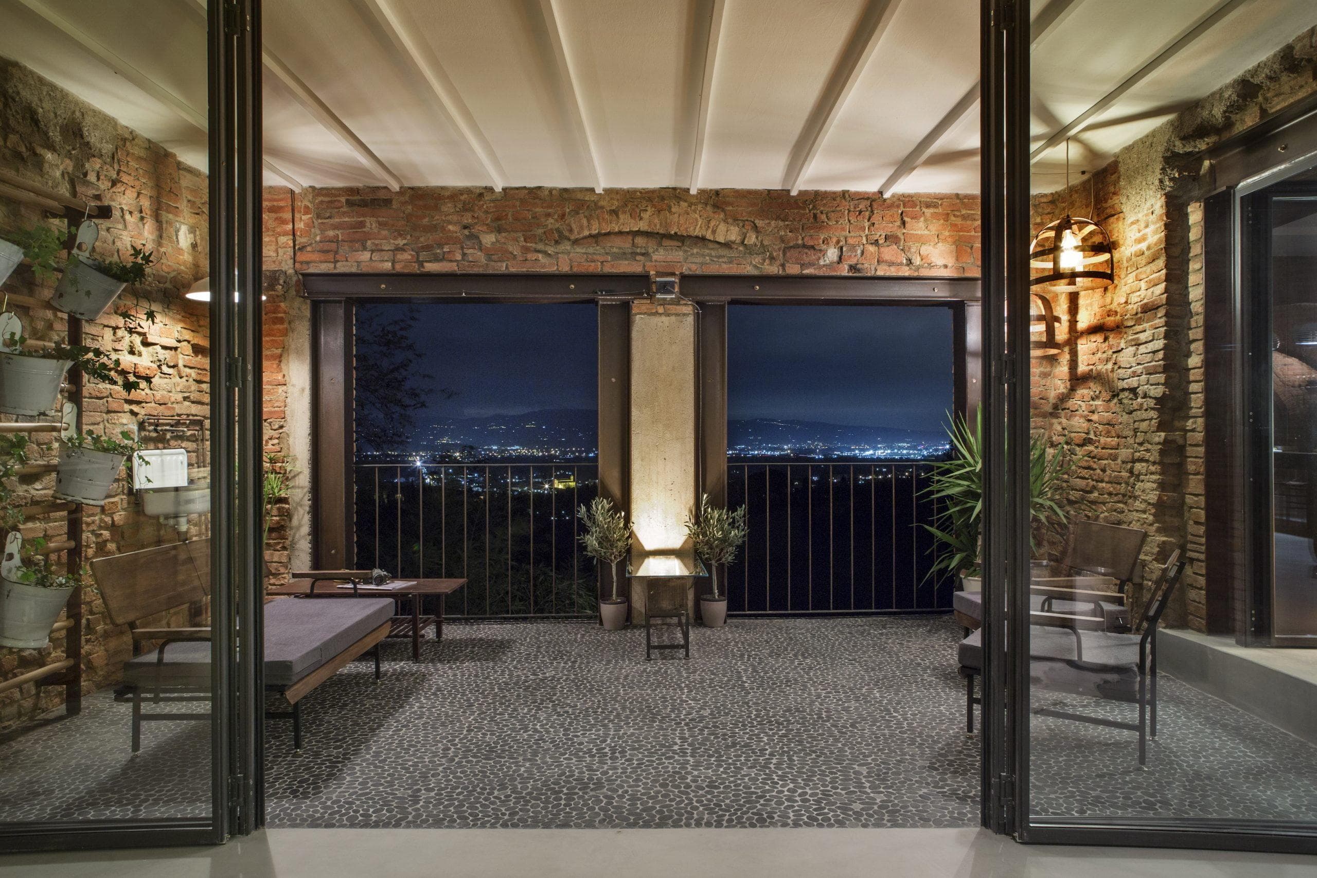 A rustic and cozy balcony with exposed brick walls, tiled flooring, and glass doors opening onto a nighttime urban landscape. The space is furnished with wooden chairs, potted plants, and a small table, all illuminated by warm lighting fixtures.