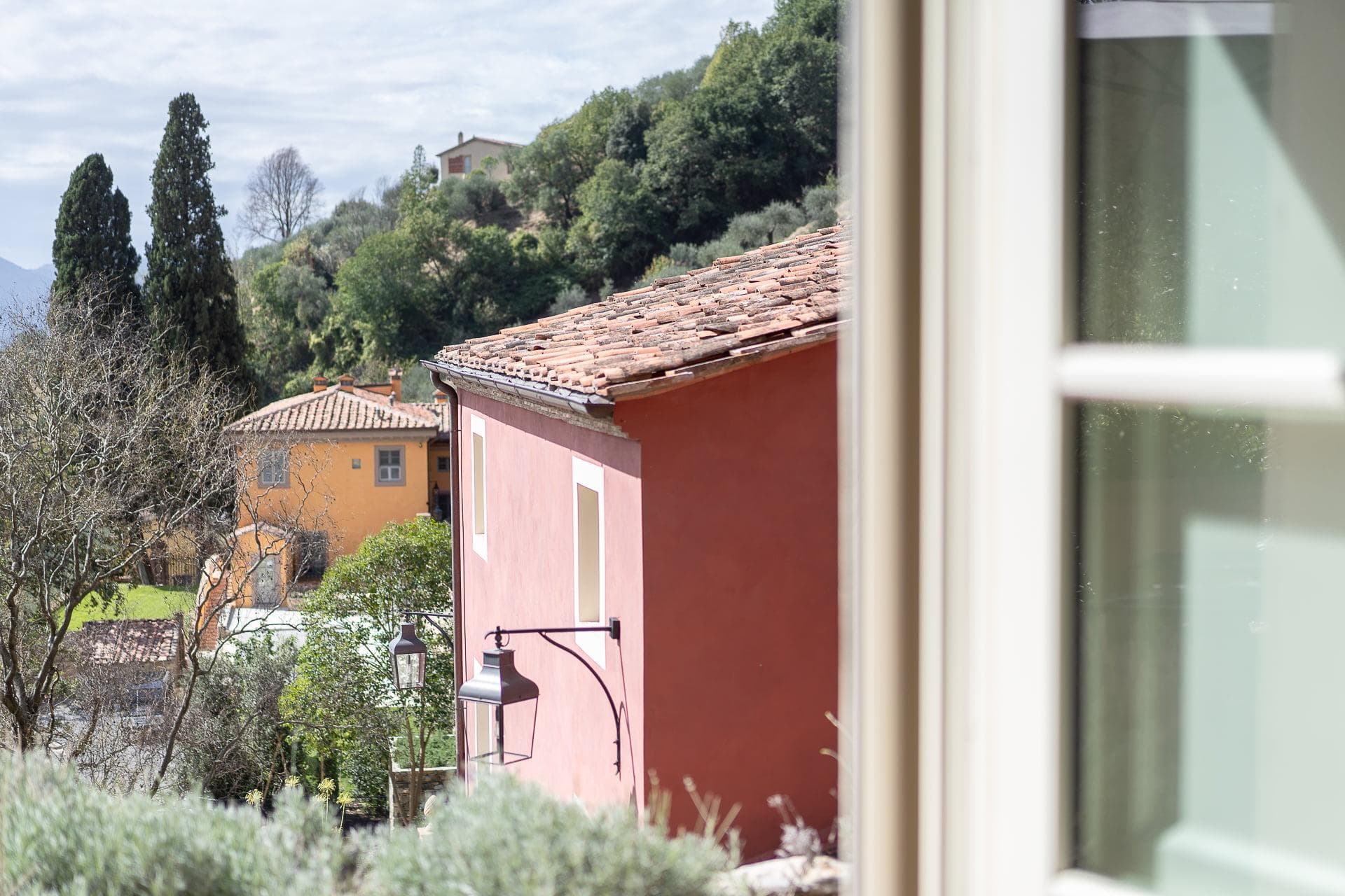 Une vue panoramique à travers une fenêtre montre des maisons aux couleurs pastel avec des toits de tuiles nichées au milieu d'une végétation luxuriante et de collines sous un ciel partiellement nuageux. Les détails du premier plan d'un cadre de fenêtre beige sont visibles sur le côté droit.