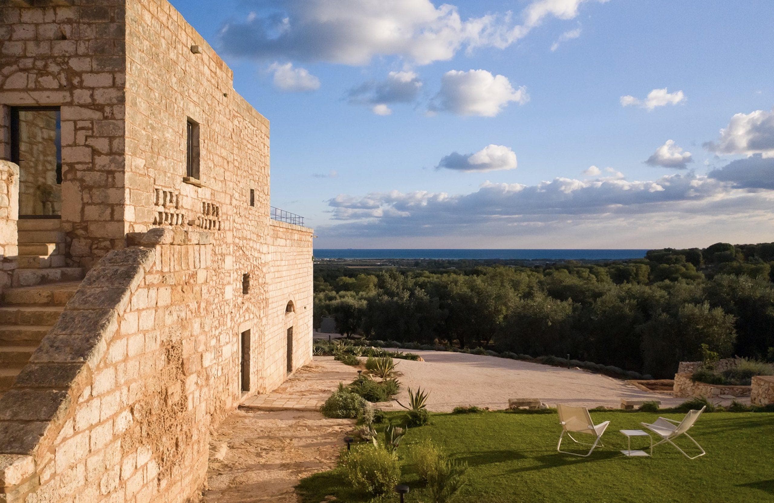 A rustic stone building with a staircase is set against a backdrop of lush greenery and olive groves, extending to a distant horizon. Two white lawn chairs are placed on a manicured lawn, with a partly cloudy sky overhead.