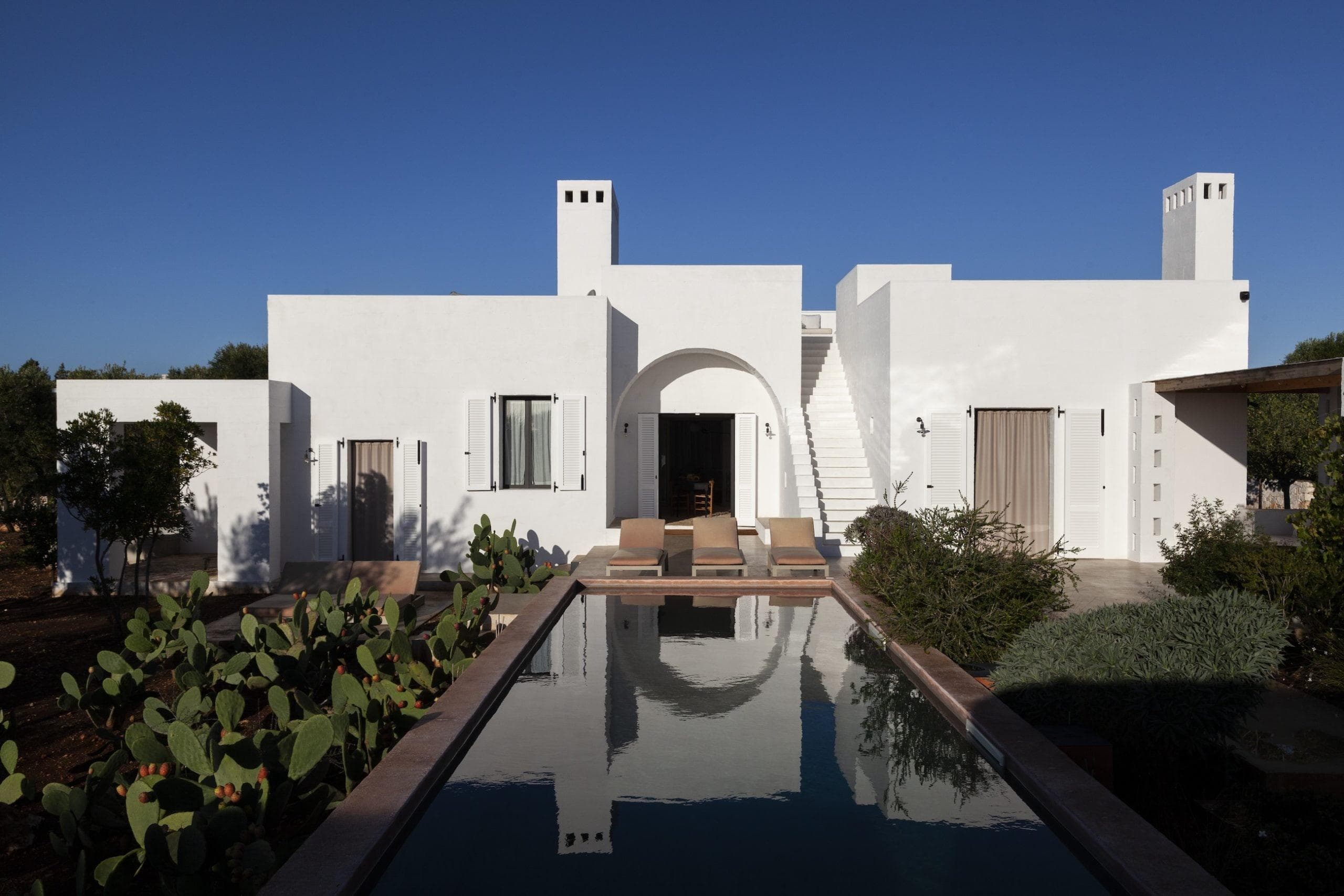 A modern white house with clean lines, featuring a central archway and flat roofs. In front, there's a rectangular swimming pool flanked by cacti and other greenery. Two lounge chairs are near the pool under a clear blue sky.