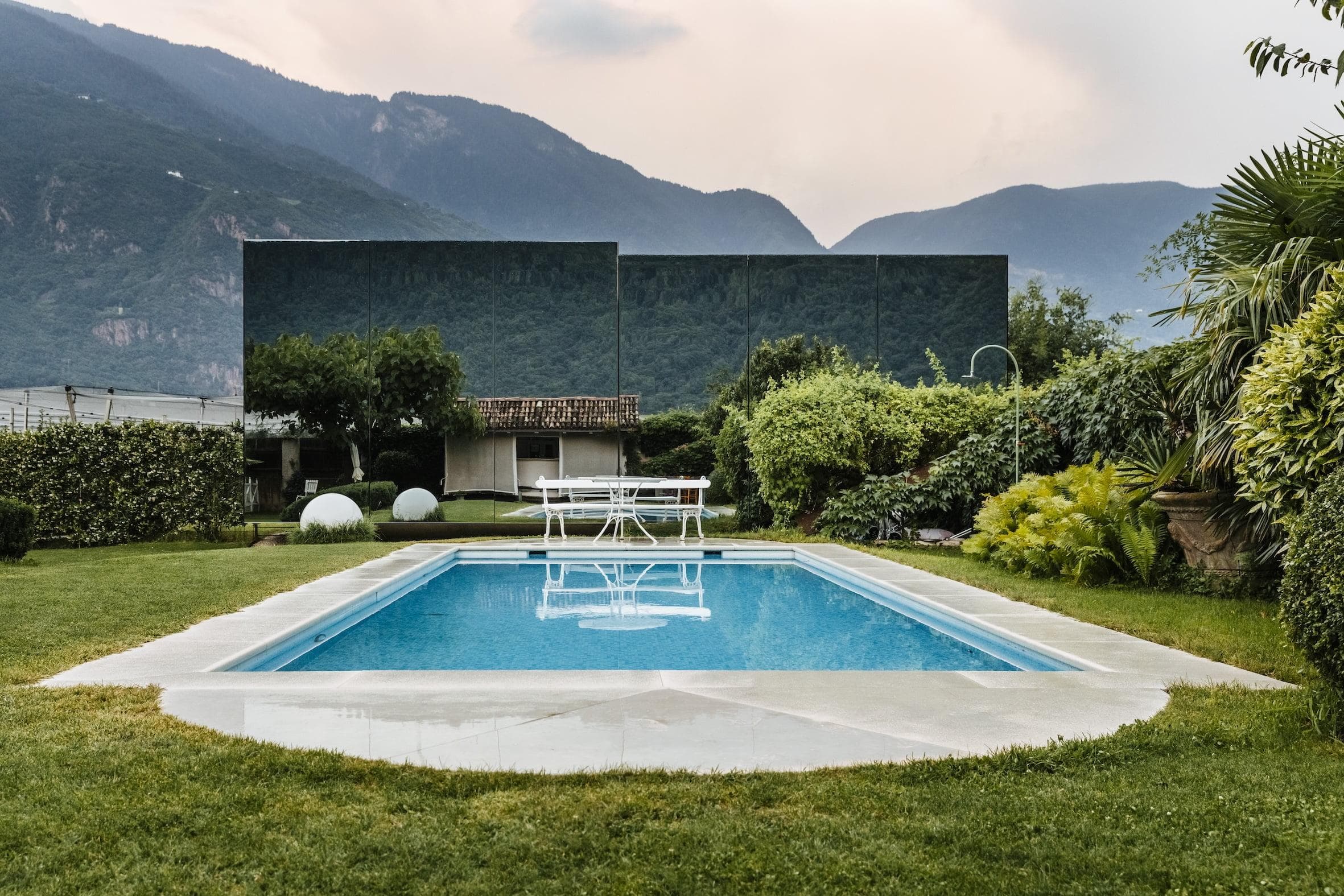 A backyard garden features a rectangular swimming pool surrounded by well-manicured grass. At the far end of the pool, there is a white outdoor dining set. Lush greenery and bushes surround the area, with mountains visible in the background under a cloudy sky.