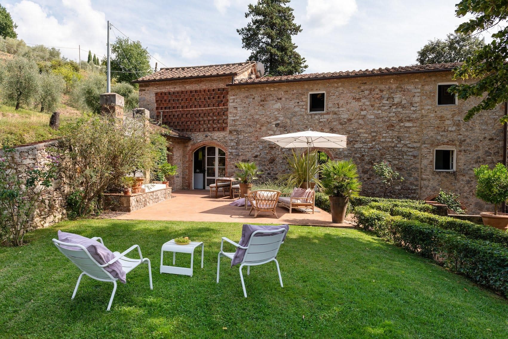 A charming stone house with a tiled roof and arched doorways sits in a lush garden. In the foreground, two white chairs and a small table with yellow flowers are set on a manicured lawn. To the right, a shaded patio features a seating area with a large umbrella.