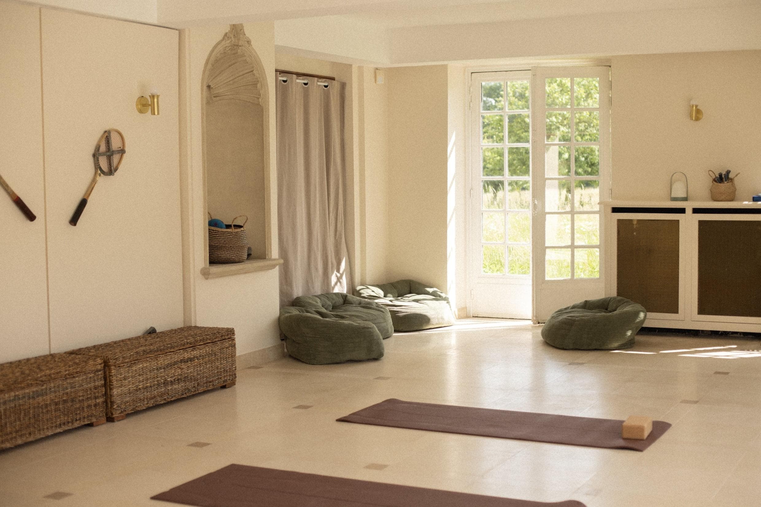 A serene yoga studio with large windows letting in natural light. The room features two yoga mats on a light-colored floor, large green cushions, and minimal decor including a wicker basket, wall-mounted tennis rackets, and a curtain.