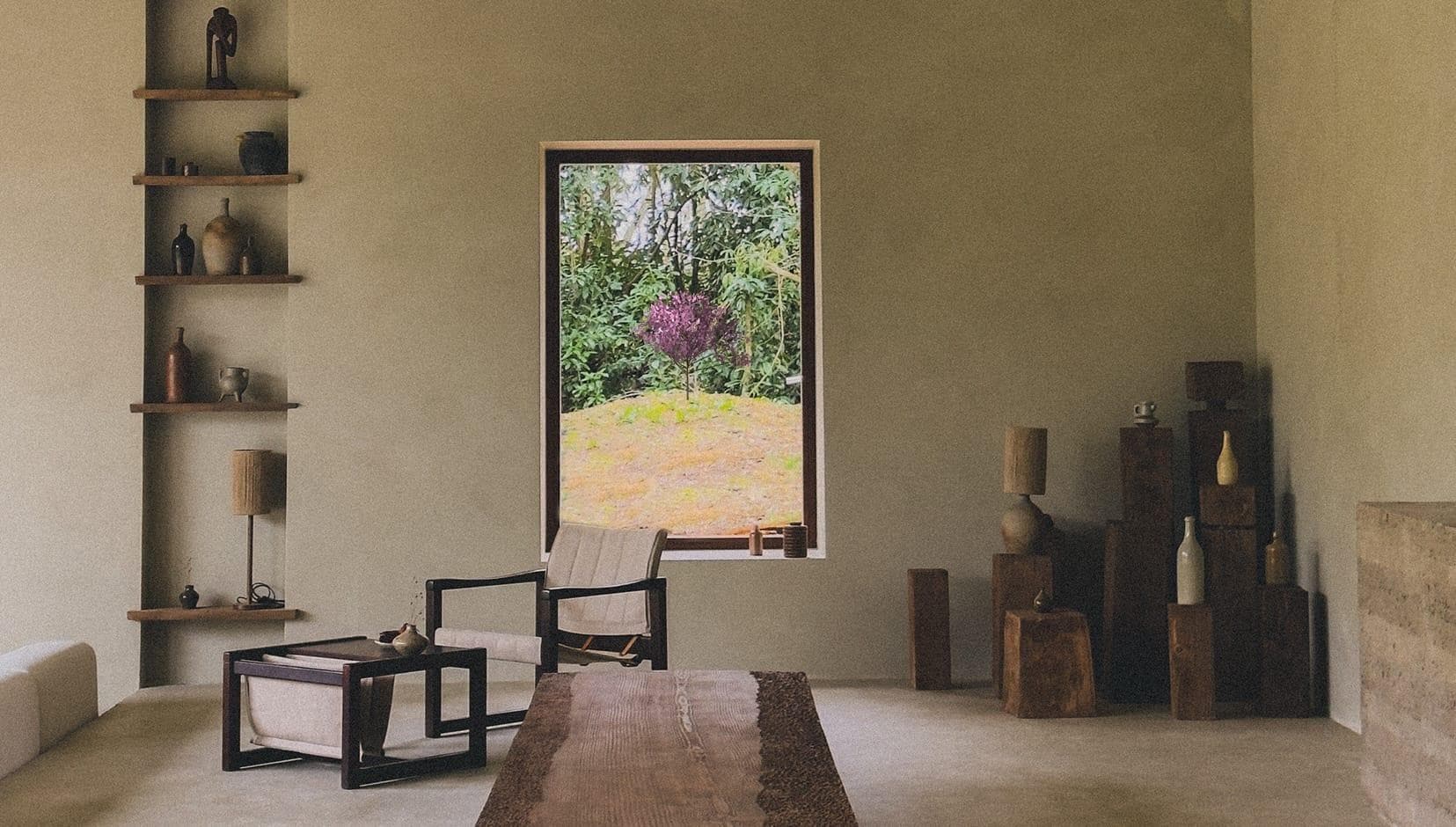Un salon minimaliste comprend un fauteuil beige, une table en bois et une décoration en bois empilé. Une grande fenêtre offre une vue sur la verdure luxuriante et un arbre violet à l'extérieur. Des tons neutres et des étagères simples avec des vases renforcent l'atmosphère sereine.