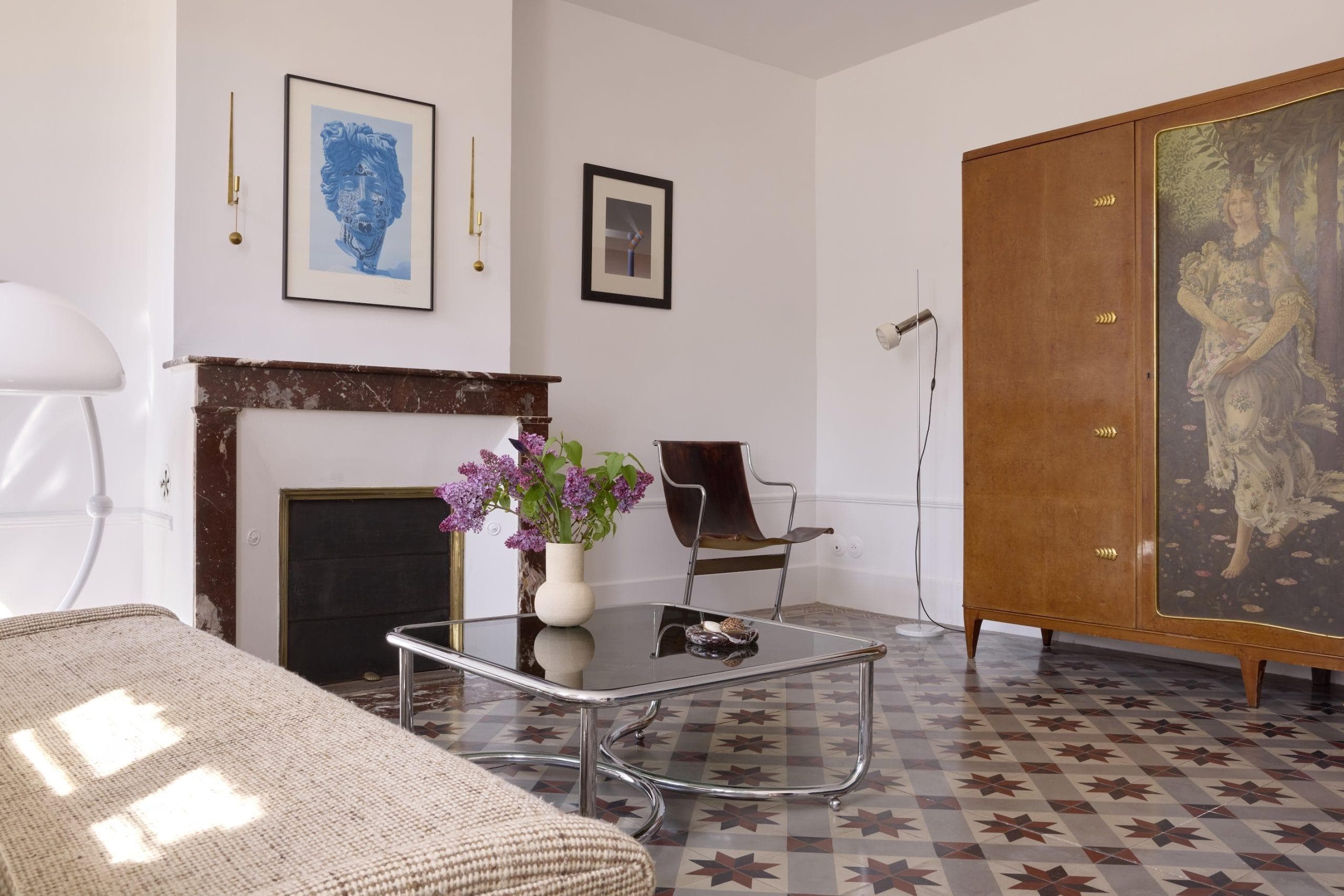 A modern living room with a geometric-patterned tile floor, featuring a beige couch, a glass coffee table with a vase of purple flowers, a wooden wardrobe, a framed artwork above the fireplace, and a floor lamp beside a leather chair.
