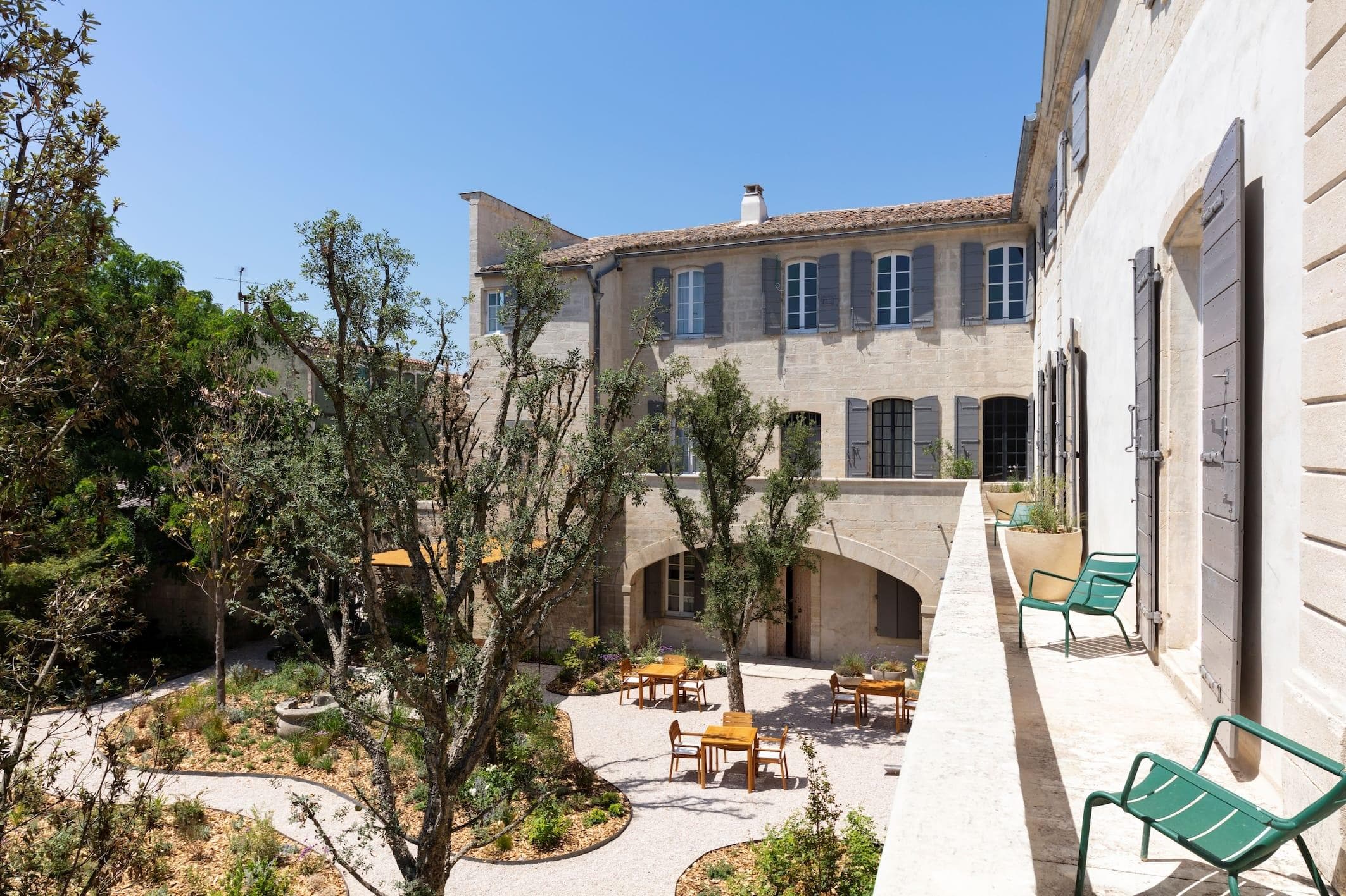 A sunny courtyard with trees and plants surrounded by a historic stone building featuring green shutters. Outdoor seating with wooden tables and chairs are scattered below a balcony with green lawn chairs.