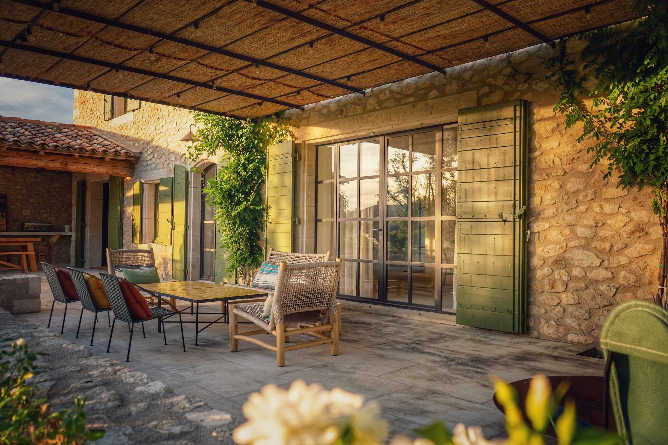 La terrasse extérieure confortable d'une maison rustique en pierre est baignée de soleil. Elle est dotée d'une table en bois entourée de chaises, de volets verts et de plantes grimpantes luxuriantes. Le premier plan montre une fleur blanche en fleurs.
