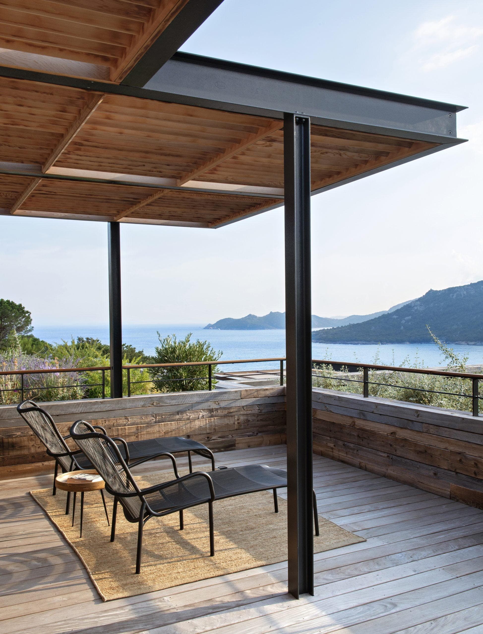 A modern outdoor terrace overlooking a scenic coastline. The terrace features two black lounge chairs on a woven rug, shaded by a wooden pergola with black metal supports. The view includes lush greenery, a tranquil sea, and distant mountains under a clear sky.