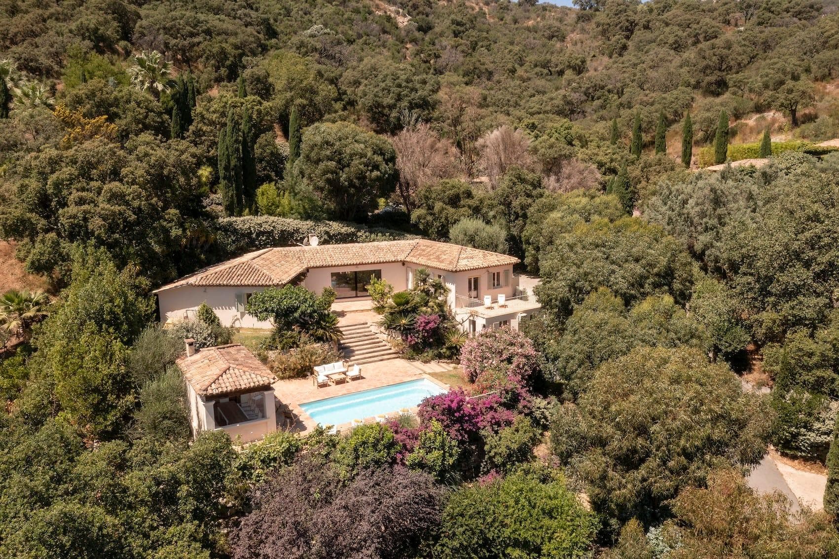 Aerial view of a villa surrounded by lush greenery and trees. The villa features a terracotta roof and a spacious patio with a rectangular swimming pool. It is situated in a hilly, forested landscape with a winding road nearby.