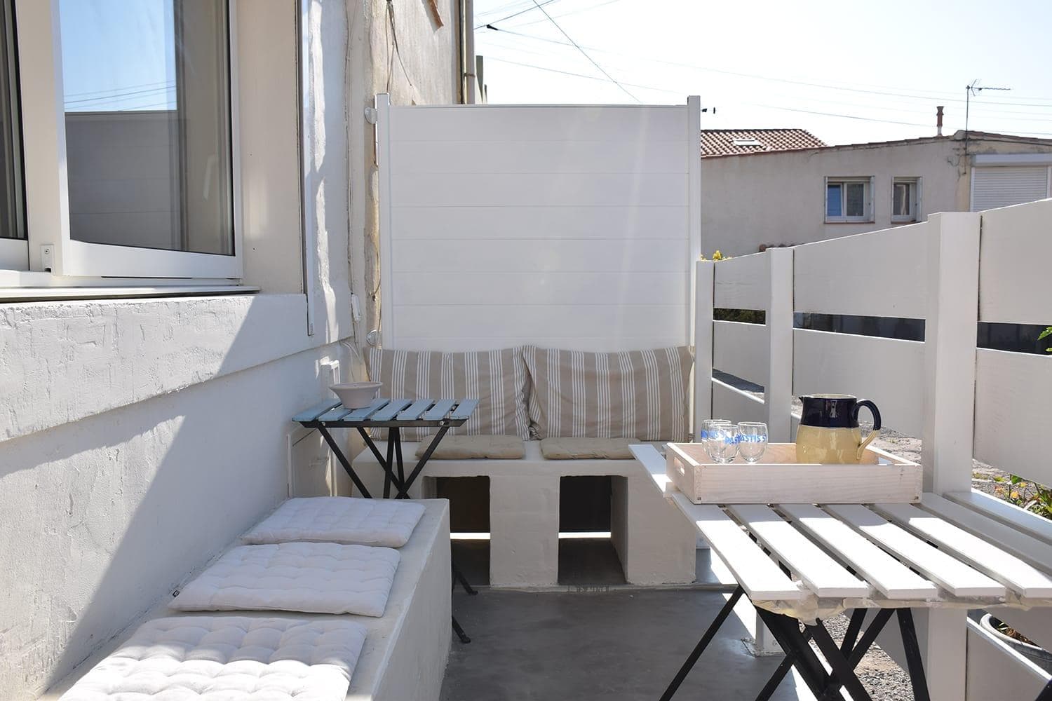 A cozy outdoor seating area featuring a cushioned bench with striped pillows against a white privacy screen. Two small tables hold a tray with a pitcher and glasses. The space is enclosed by white fencing, with sunlight casting soft shadows.
