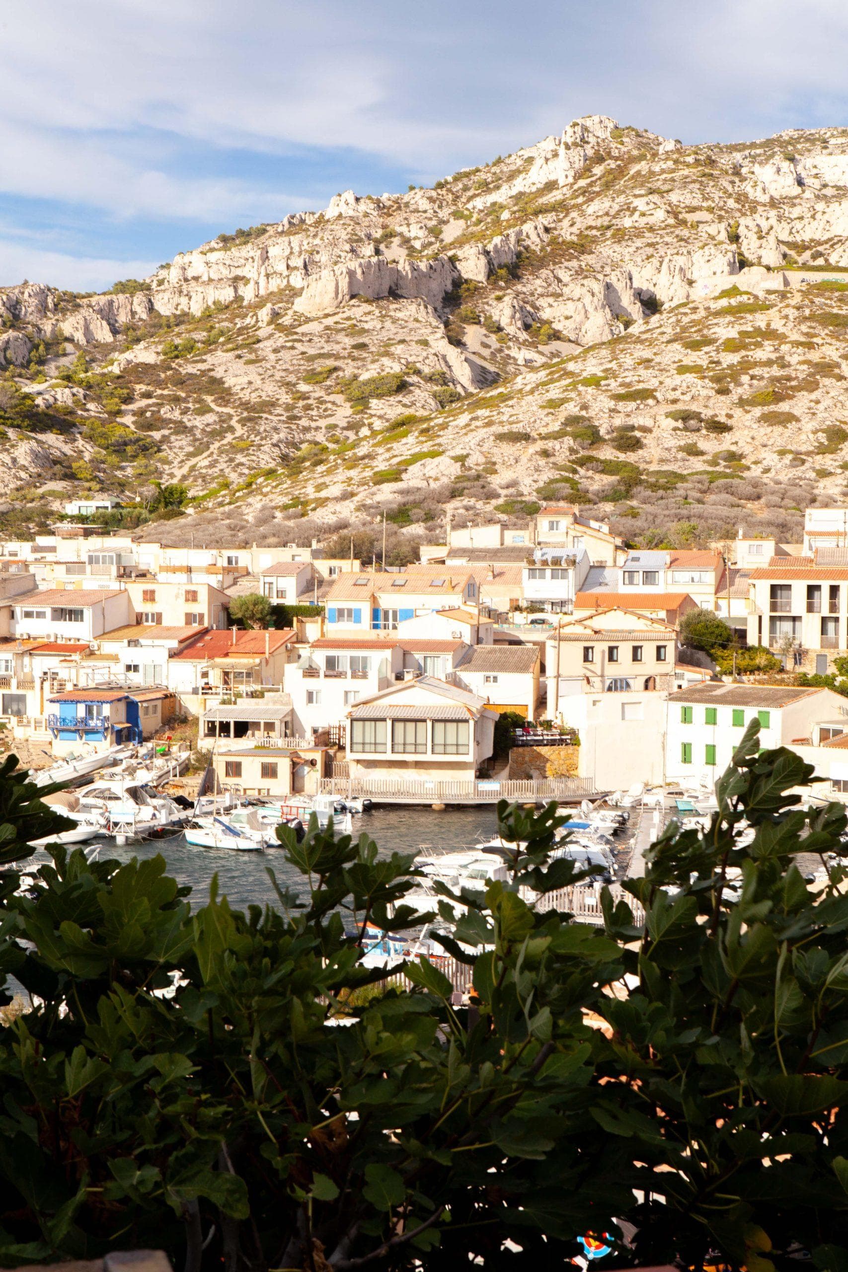 Vue d'une ville portuaire pittoresque nichée contre des collines rocheuses et escarpées. La scène présente de nombreuses maisons serrées avec des couleurs et des styles variés, un front de mer calme avec des bateaux amarrés et une verdure luxuriante au premier plan. Le ciel est partiellement nuageux.