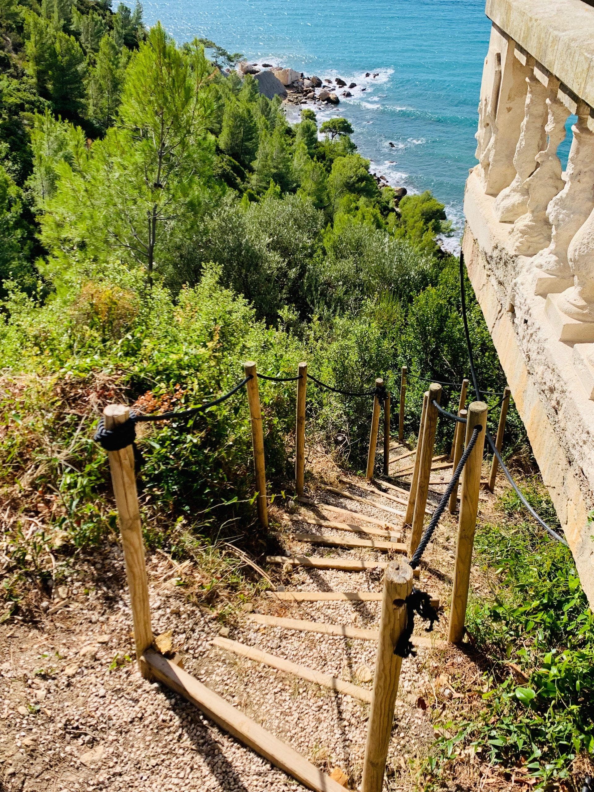 A narrow wooden staircase descends steeply down a grassy hill, bordered by a stone railing. In the background, the panoramic view of this luxurious location reveals the sea in a vibrant blue, meeting a rocky coastline on the French Riviera, all framed by lush green trees.