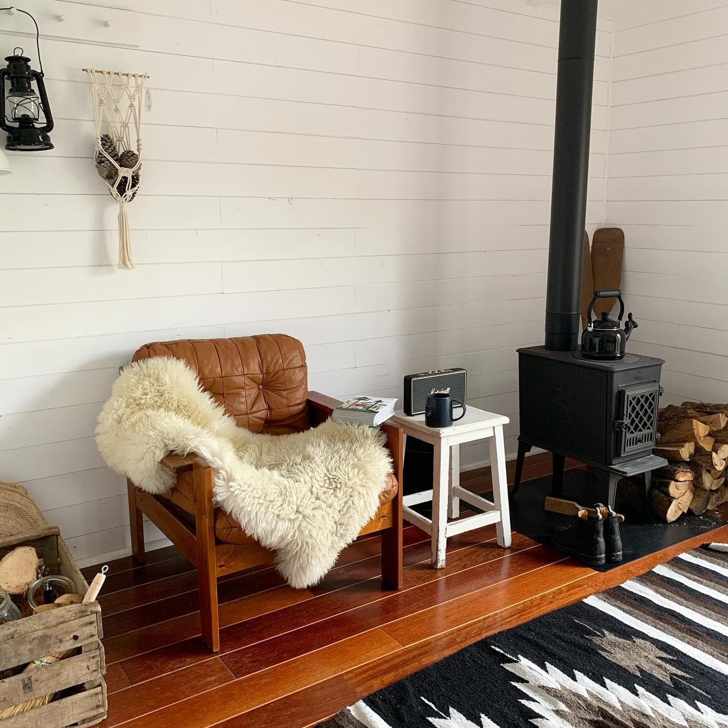 Une pièce confortable avec un fauteuil en cuir recouvert d'un tapis en peau de mouton, un petit tabouret blanc, un poêle à bois avec une bouilloire, une pile de bois de chauffage et un panier. Le sol est en bois avec un tapis à motifs et un macramé suspendu décore le mur blanc.