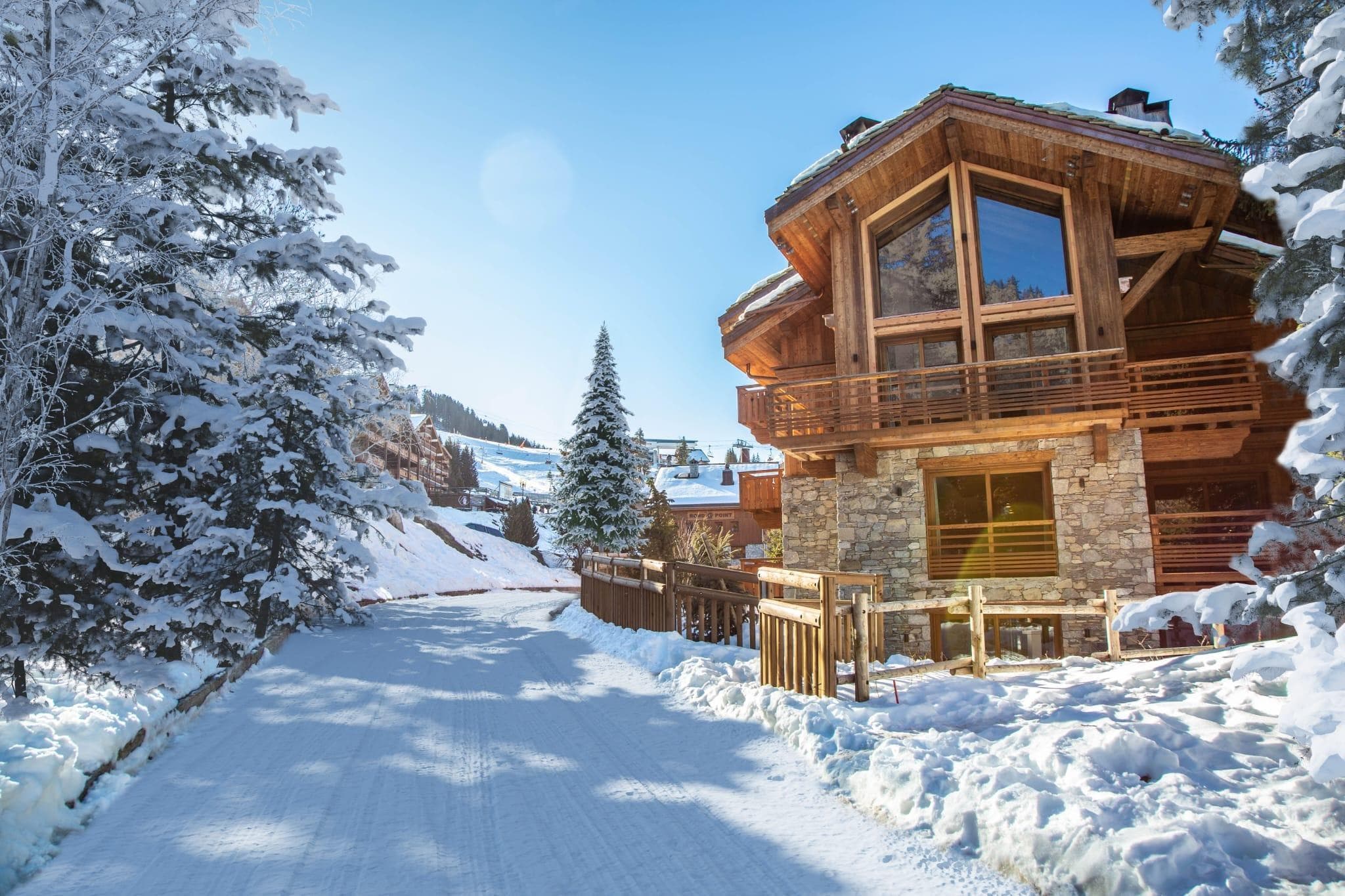 A wooden chalet with large windows and a stone base is surrounded by snow-covered trees on a sunny day. A snow-covered path leads to the chalet, and mountains are visible in the background.