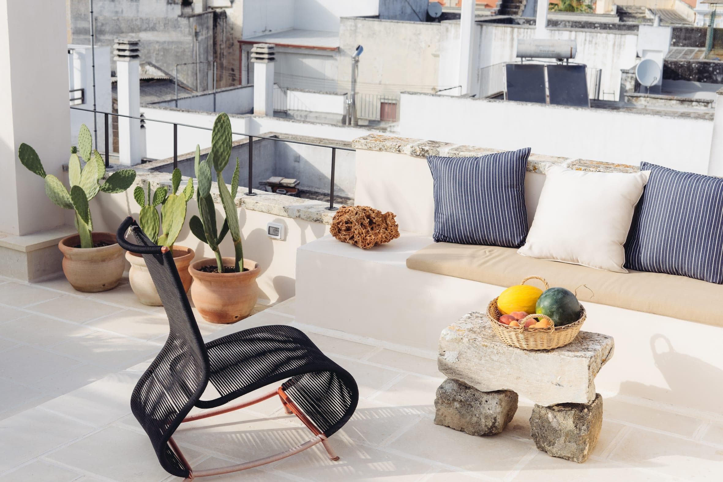 A rooftop patio features a modern black chair and a stone table with a basket of fruit. Cushioned seating lines the wall, and potted cacti add greenery. The background shows a cityscape with buildings under a clear sky.