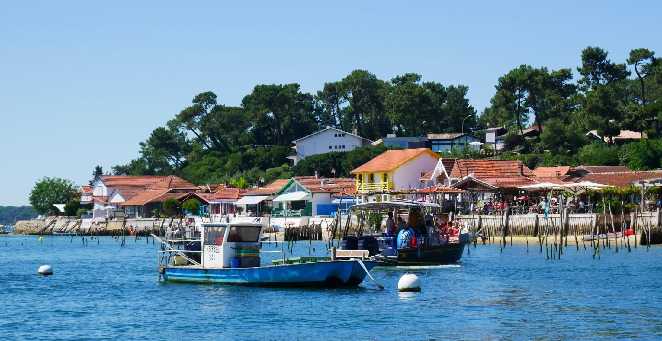 A serene coastal village with colorful houses lined along the shore. Several small boats are anchored in the blue water, and a lush green forest rises behind the buildings. People are visible on the waterfront, enjoying the sunny day.