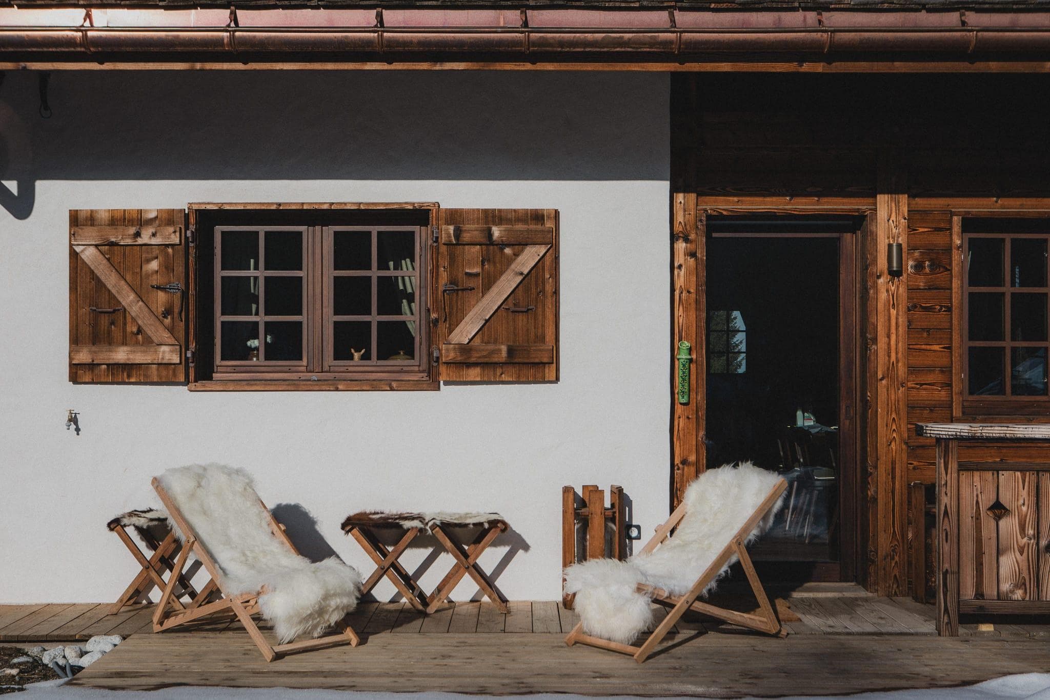 Two wooden deck chairs with white fur throws face each other on a sunlit wooden porch. A small table sits between them. The exterior features rustic wooden shutters and a partially open door leading inside.