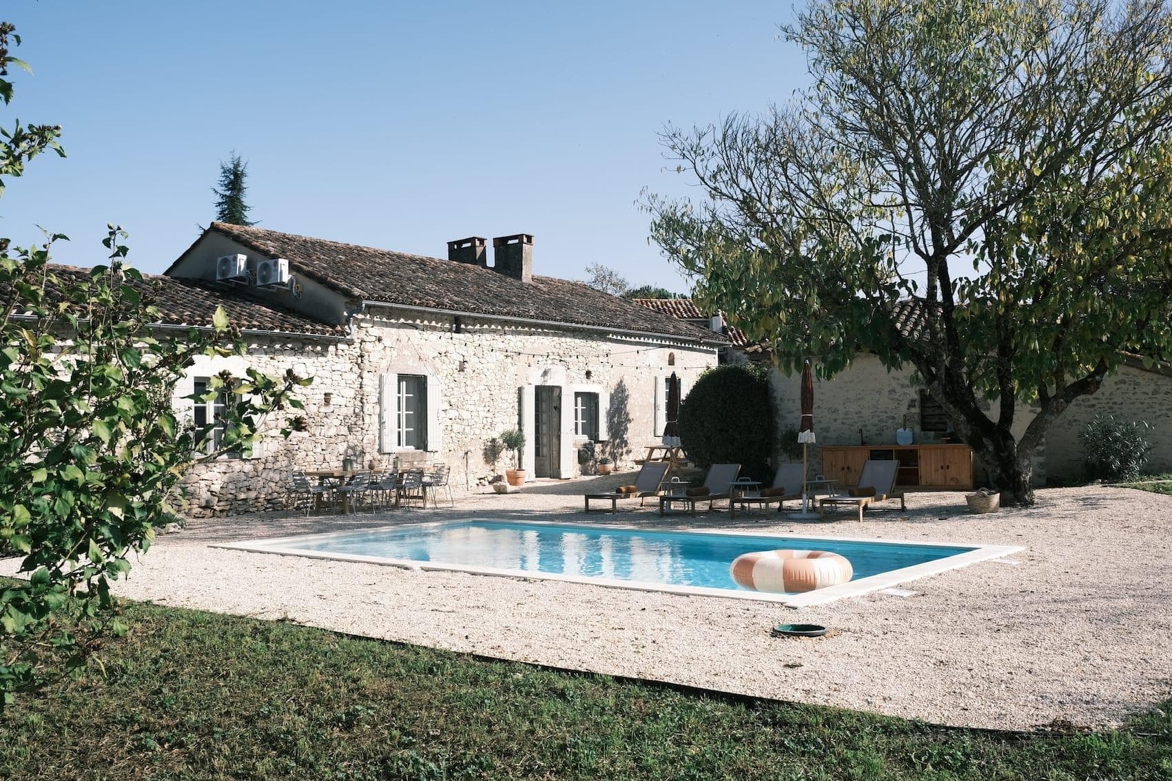 A stone house with a tiled roof and green shutters stands next to a light-blue swimming pool. Loungers and a tree create a relaxed atmosphere in the garden. The sky is clear and blue, reinforcing the tranquil setting.