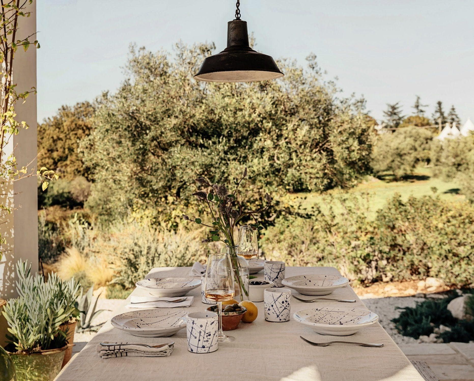 A rustic outdoor dining table is set with white patterned plates, bowls, and glasses, topped with simple decor including a vase with flowers. Overhead, a single black lamp hangs. The background shows a lush green garden with trees under a clear sky.