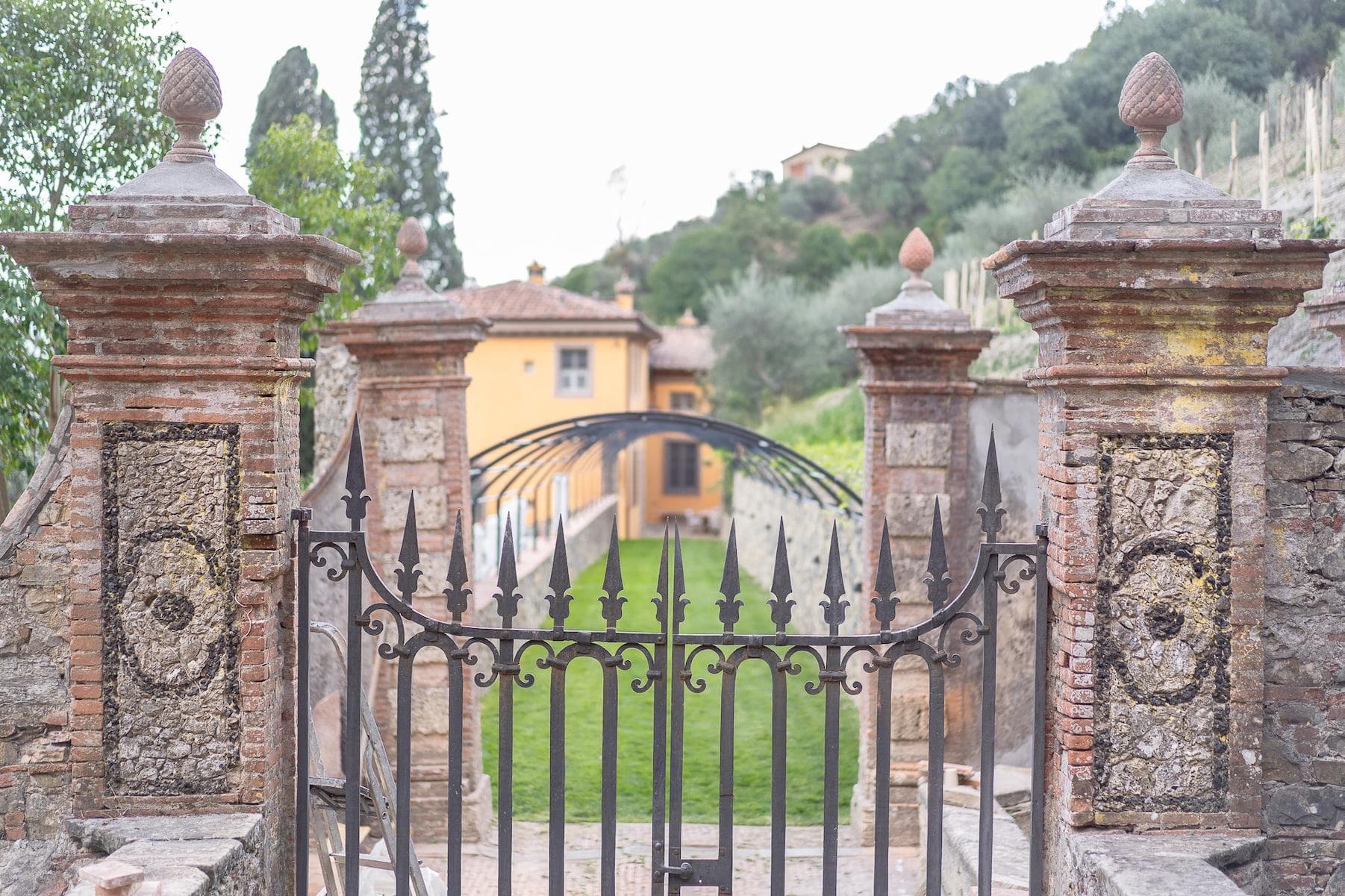 A wrought-iron gate with intricately designed stone pillars opens onto a pathway flanked by green lawns and garden arches. A yellow building with a tiled roof is visible in the background, set against a backdrop of trees and lush vegetation.