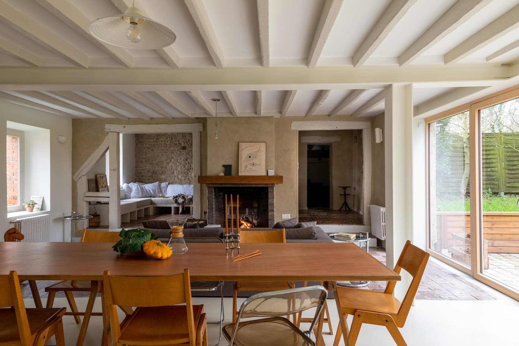 A cozy living-dining room, adorned with wooden beams, features a long table surrounded by mismatched chairs, a lit fireplace and a sofa. Large windows let in natural light, and decorations include a pumpkin and candlesticks on the table.