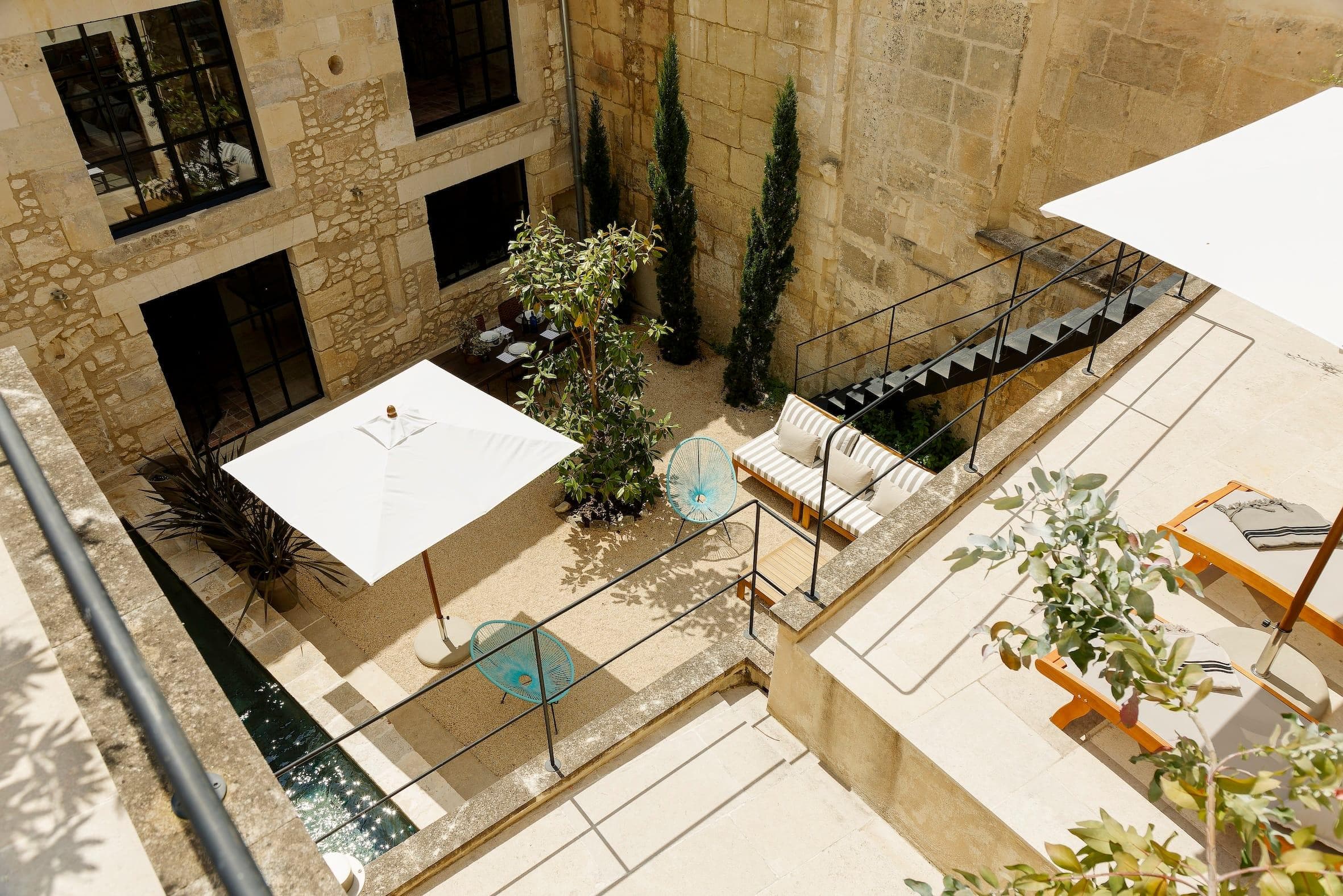 A sunny stone courtyard features a white parasol above a seating area with striped cushions, small round tables, green plants and deckchairs next to a narrow reflecting pool
