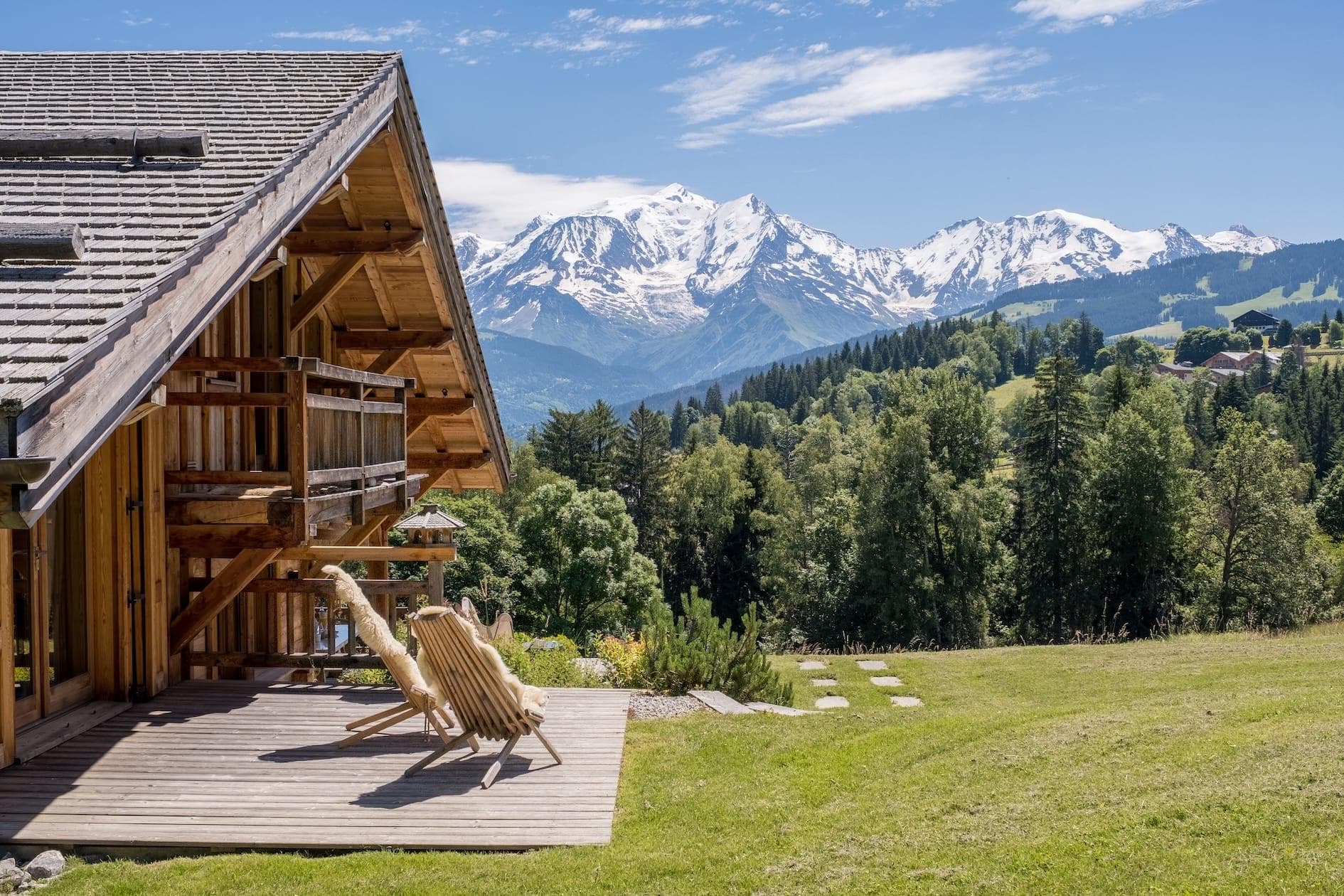 A wooden cabin with a porch featuring two lounge chairs overlooks a scenic mountain range with snow-capped peaks and lush green forest. The sky is clear with a few clouds, enhancing the natural beauty of the landscape.
