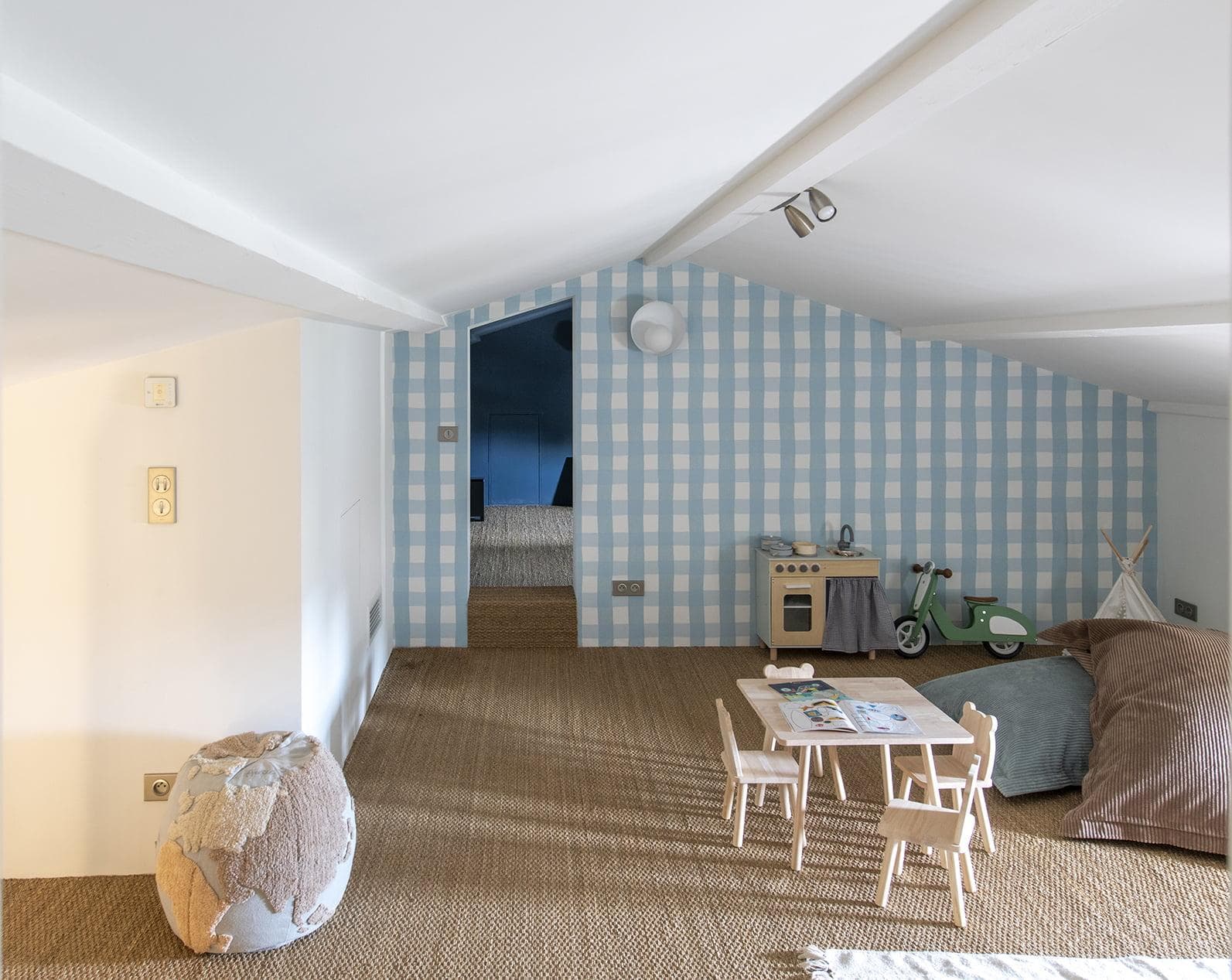 A cozy children's playroom with sloped ceilings, blue gingham wallpaper, a small wooden table and chairs, a toy kitchen, a green scooter, a teepee, large floor cushions, and a globe-shaped pouf.
