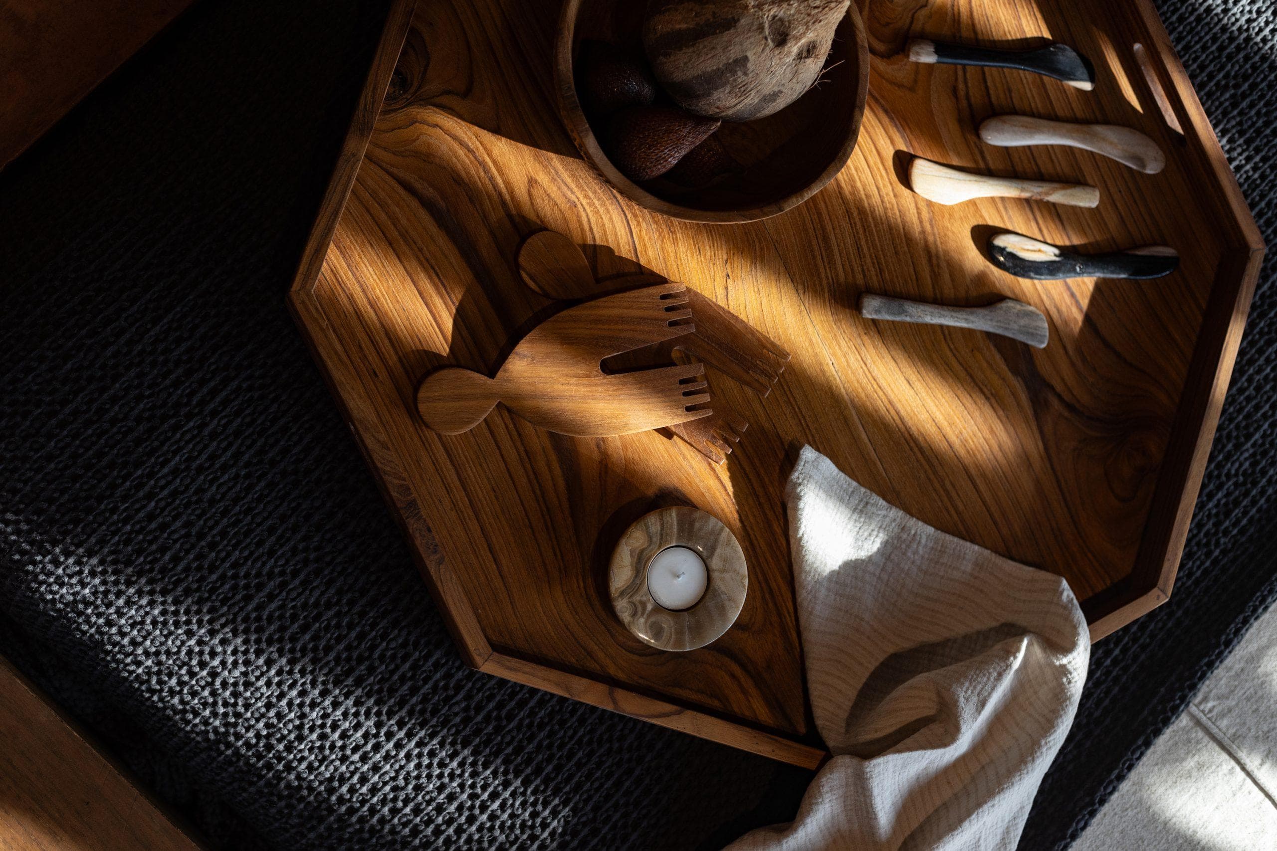 Un plateau en bois repose sur une surface texturée sombre, éclairée par la lumière naturelle. Sur le plateau se trouvent plusieurs objets dont trois petits couteaux aux manches légers, un objet en bois en forme de poisson, un bol et une petite bougie à côté d'un tissu blanc plié.