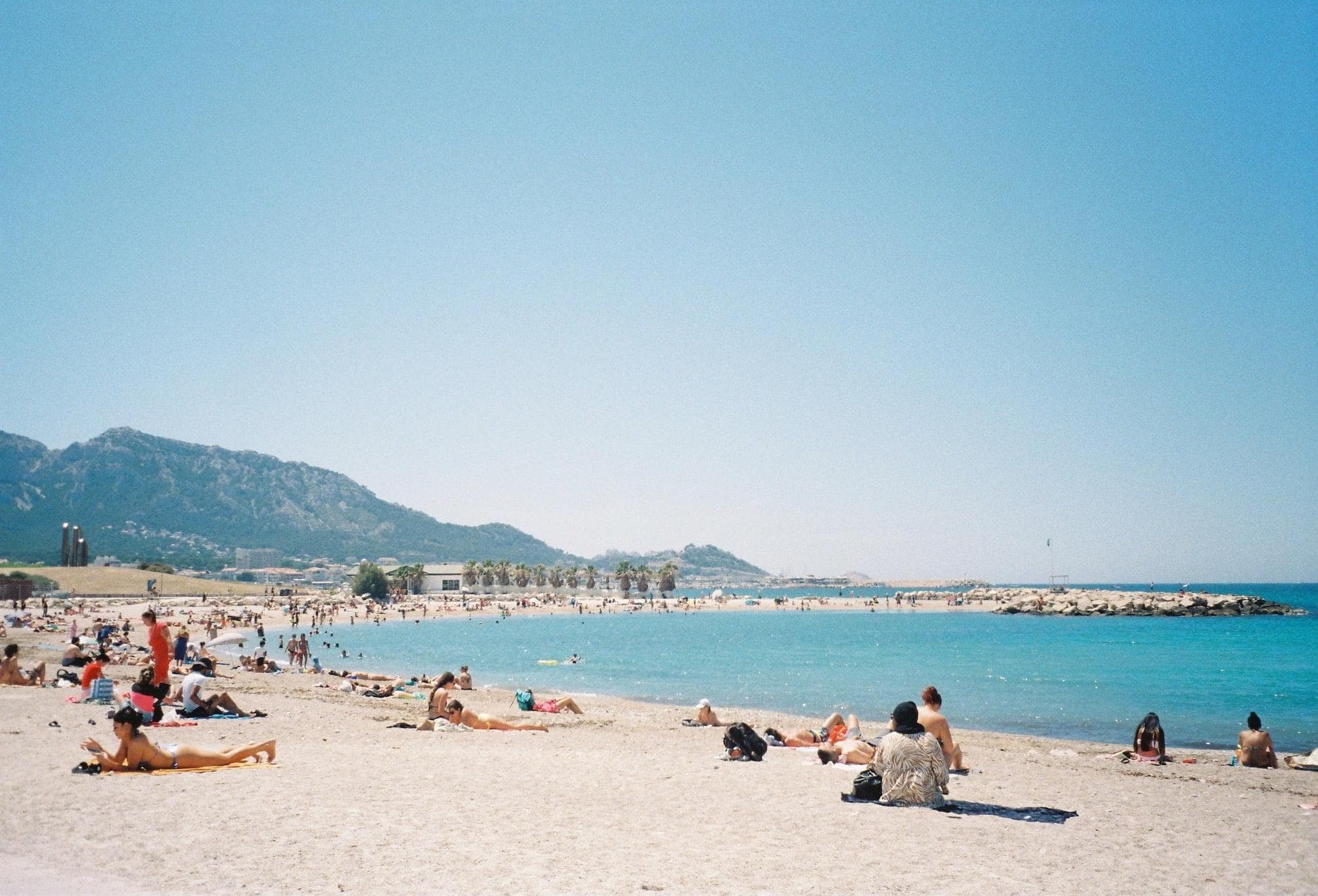 Une scène de plage ensoleillée avec des gens bronzant sur le rivage sablonneux et nageant dans la mer turquoise. En arrière-plan, se dressent une chaîne de collines, partiellement recouvertes de verdure. Le ciel clair ajoute à l’atmosphère sereine et lumineuse.