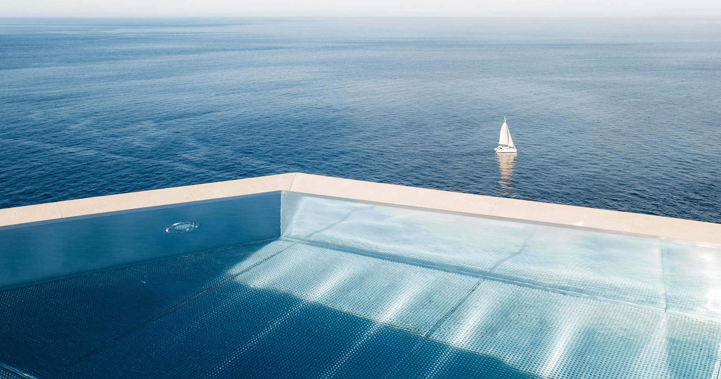An infinity pool with a clear blue surface, adjoining a vast and tranquil ocean. A single white sailboat is visible in the distance on the horizon, under a clear sky. The entire scene exudes calmness and serenity.