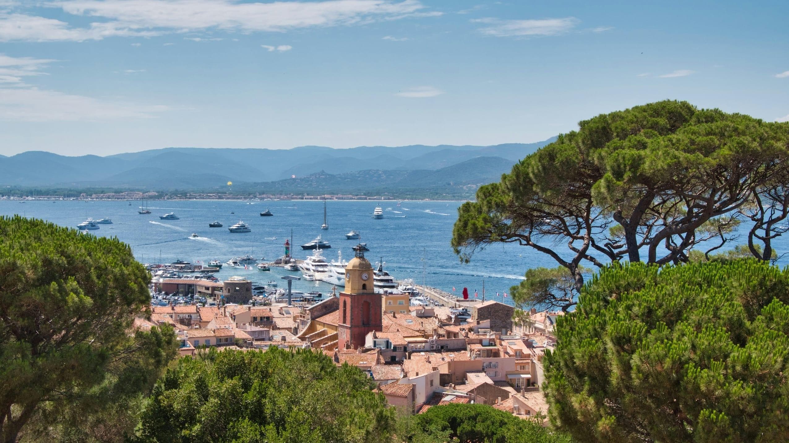 Vue d'une ville côtière près de Saint-Tropez, avec des toits en terre cuite et un clocher d'église proéminent. Une marina animée prospère tandis que la mer est parsemée de bateaux. Des collines vallonnées et des arbres luxuriants encadrent cet endroit idyllique sous un ciel bleu clair, parfait pour découvrir votre escapade dans une villa de rêve.