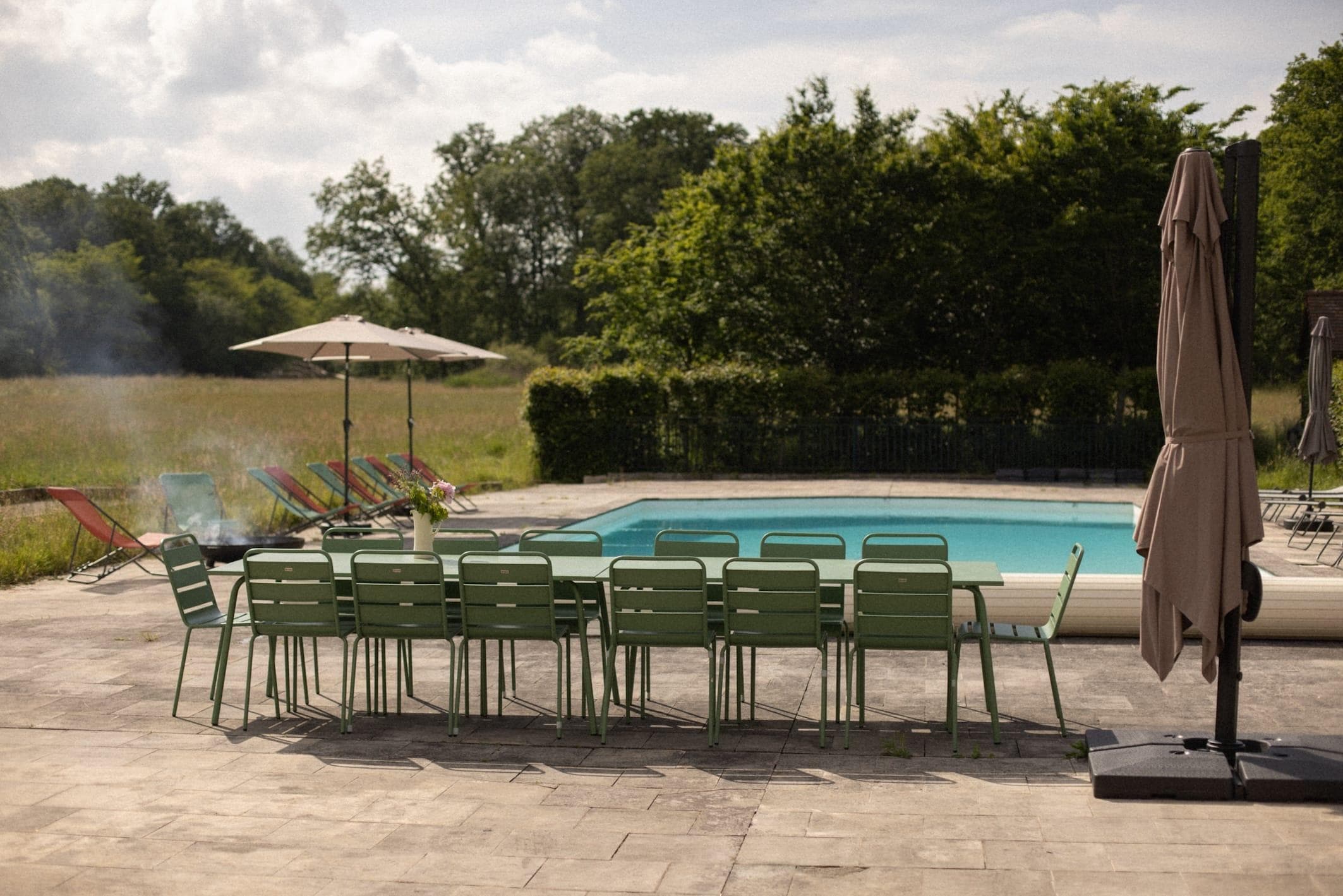 Une longue table d'extérieur et des chaises vertes sont installées sur une terrasse en pierre au bord d'une piscine, évoquant le charme d'une grande maison de vacances. Des chaises longues et des parasols sont disposés à proximité, entourés d'arbres et de pelouse, sous un ciel partiellement nuageux.