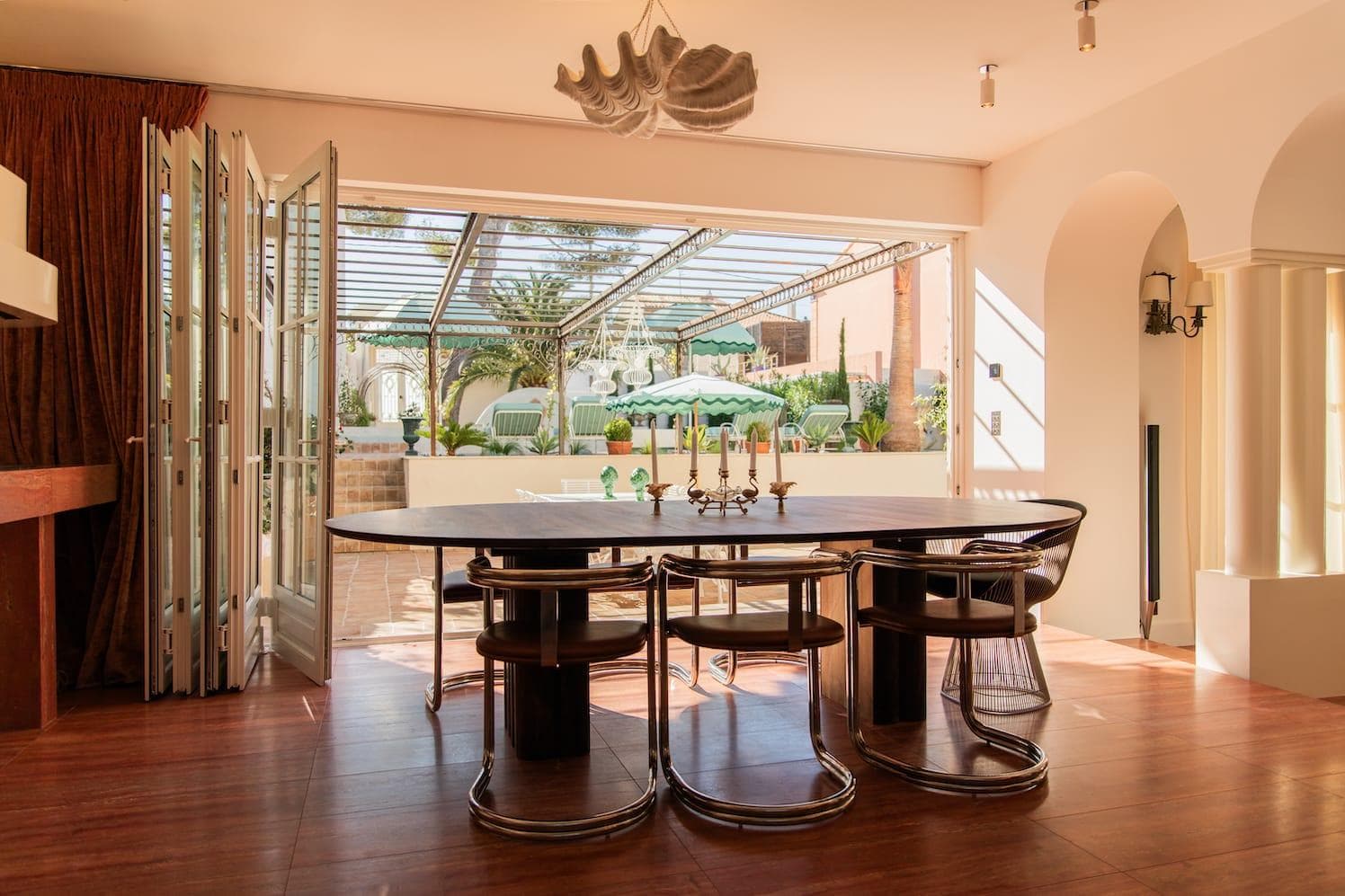 A modern dining room with a large dark oval table and eight chairs, open folding patio doors open onto a sunny terrace with plants, seating and a green-striped parasol. A sculptural suspension hangs over the table.