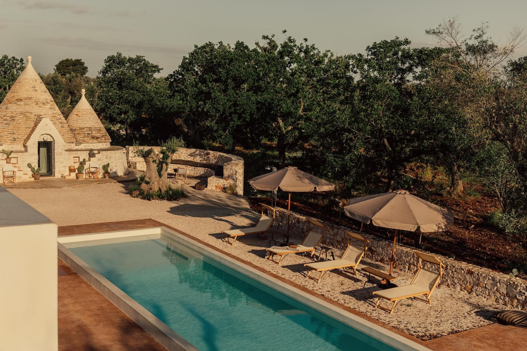A rectangular swimming pool with lounge chairs and umbrellas is set beside a traditional stone trullo house, surrounded by trees and rustic stone walls under a clear sky.