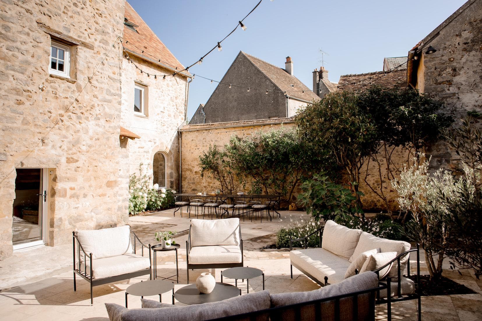 A sunlit stone courtyard with cream outdoor sofas, round black tables, and string lights overhead. Green plants line the walls, and old stone buildings surround the cozy seating area.